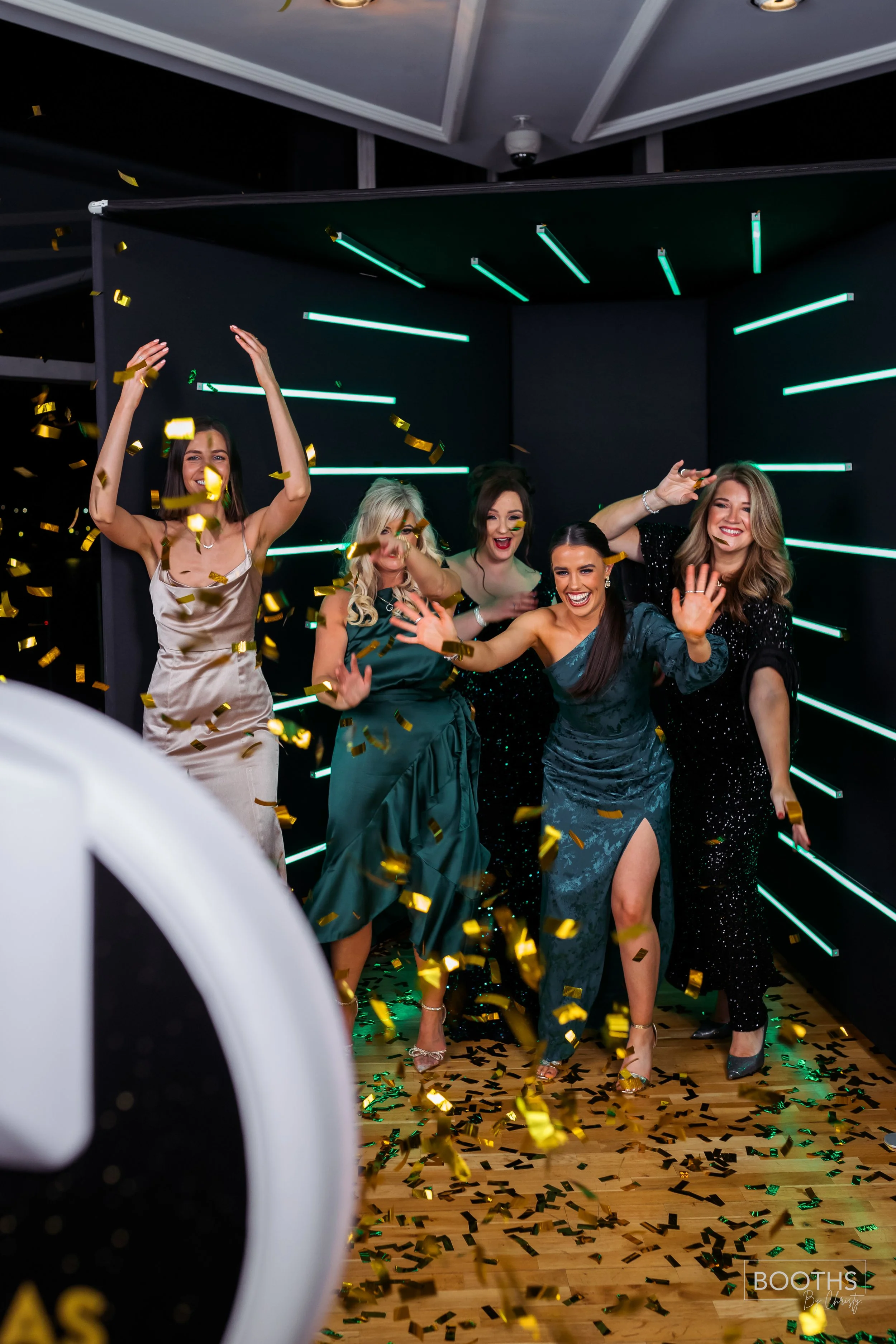 Group of women celebrating with gold confetti, smiling, dancing, dressed in evening gowns, in a decorated indoor venue