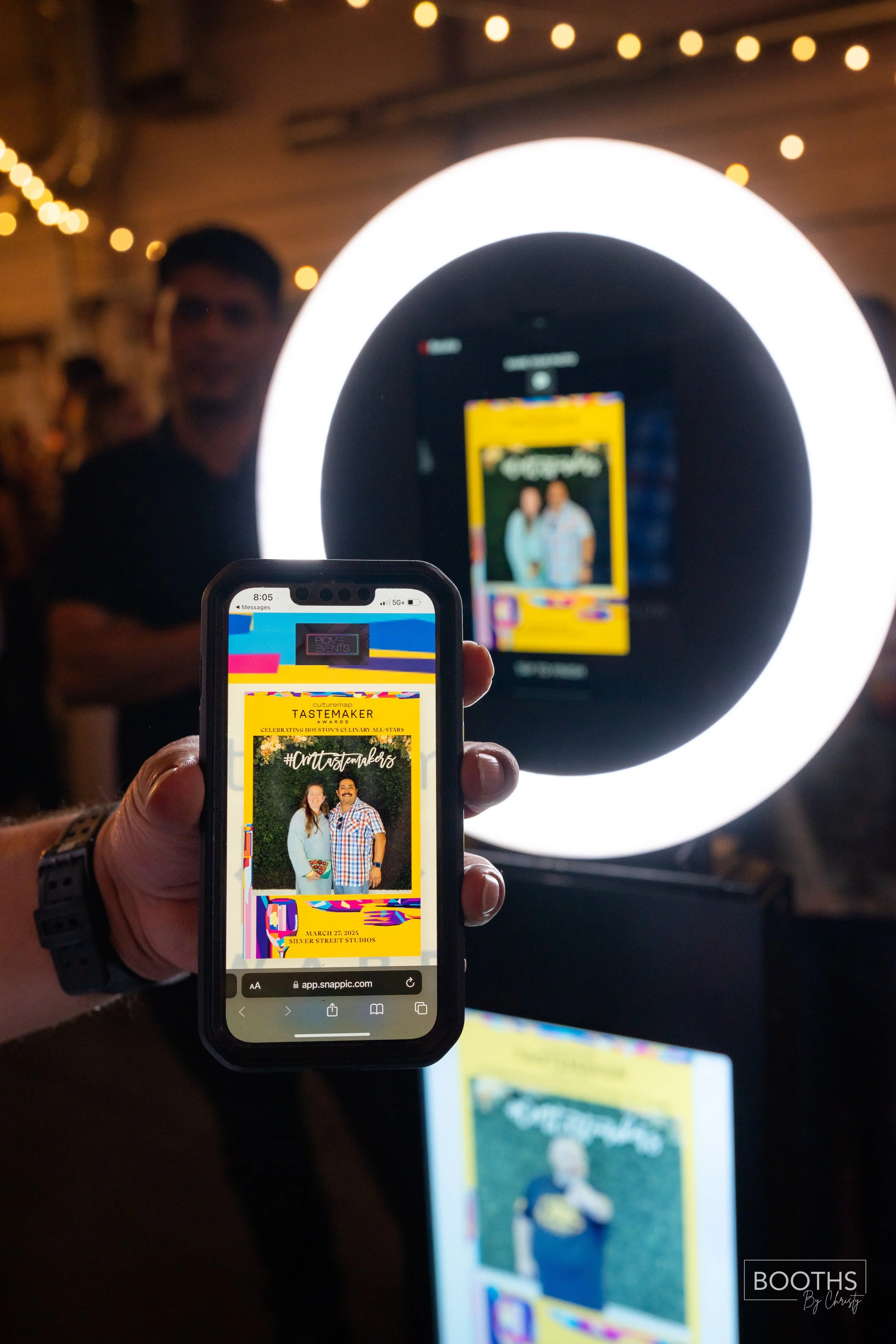 Person holding a smartphone with a digital event ticket displayed, featuring two people on a photo, with a large ring light and a blurred background of people at an indoor event.