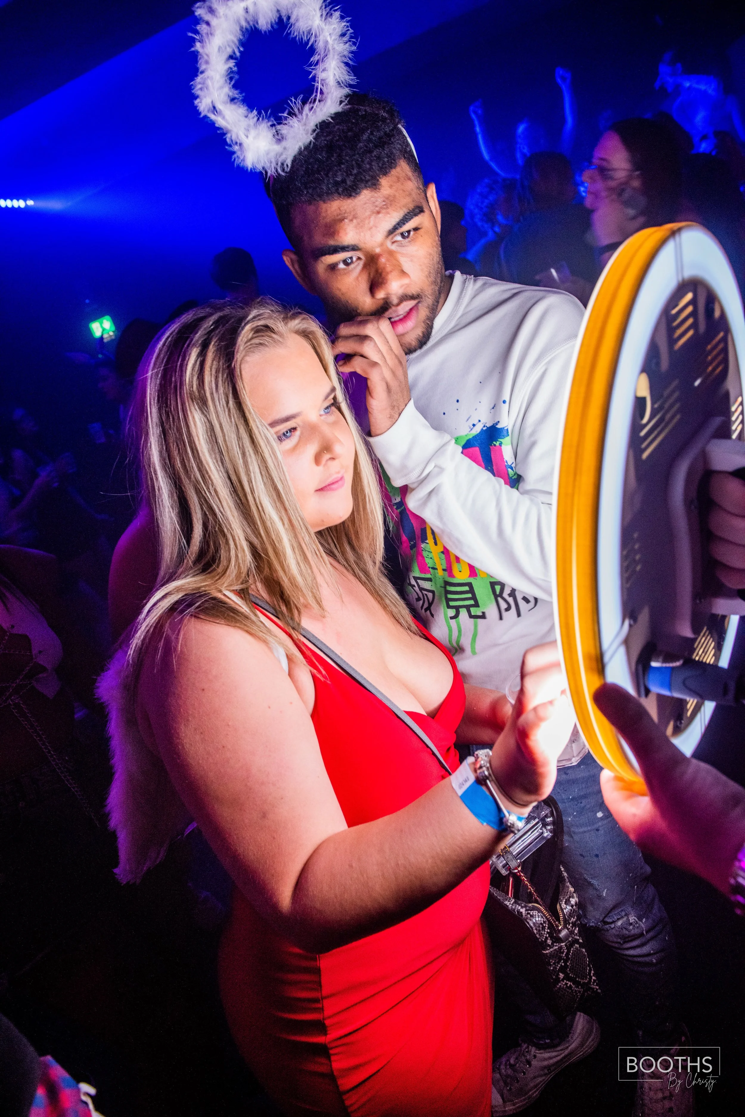 A woman in a red dress and a man with angel wings headband taking a selfie at a party, with colorful lighting and other guests in the background.