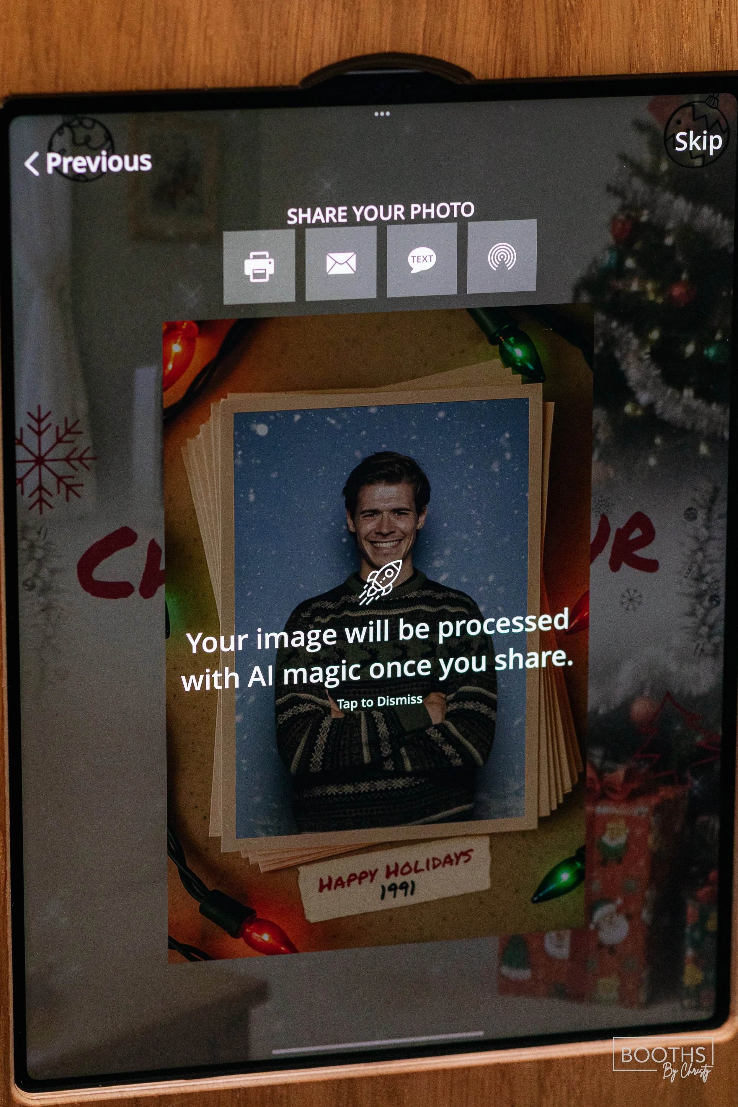 A smartphone screen displaying a photo of a smiling young man with dark hair, wearing a striped sweater, standing in snowfall. The photo is on top of holiday cards and decorated with Christmas lights and decorations.