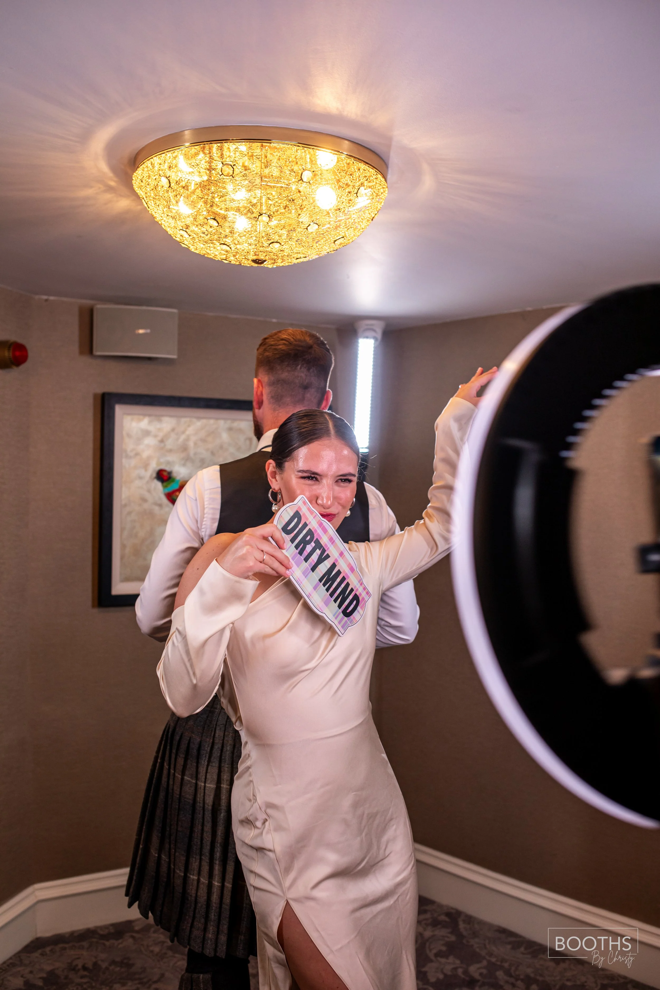 A woman in a cream-colored dress holding a sign that says 'DIRTY MIND' while dancing in a room with a man dressed in formal attire behind her, with a decorative ceiling light overhead.