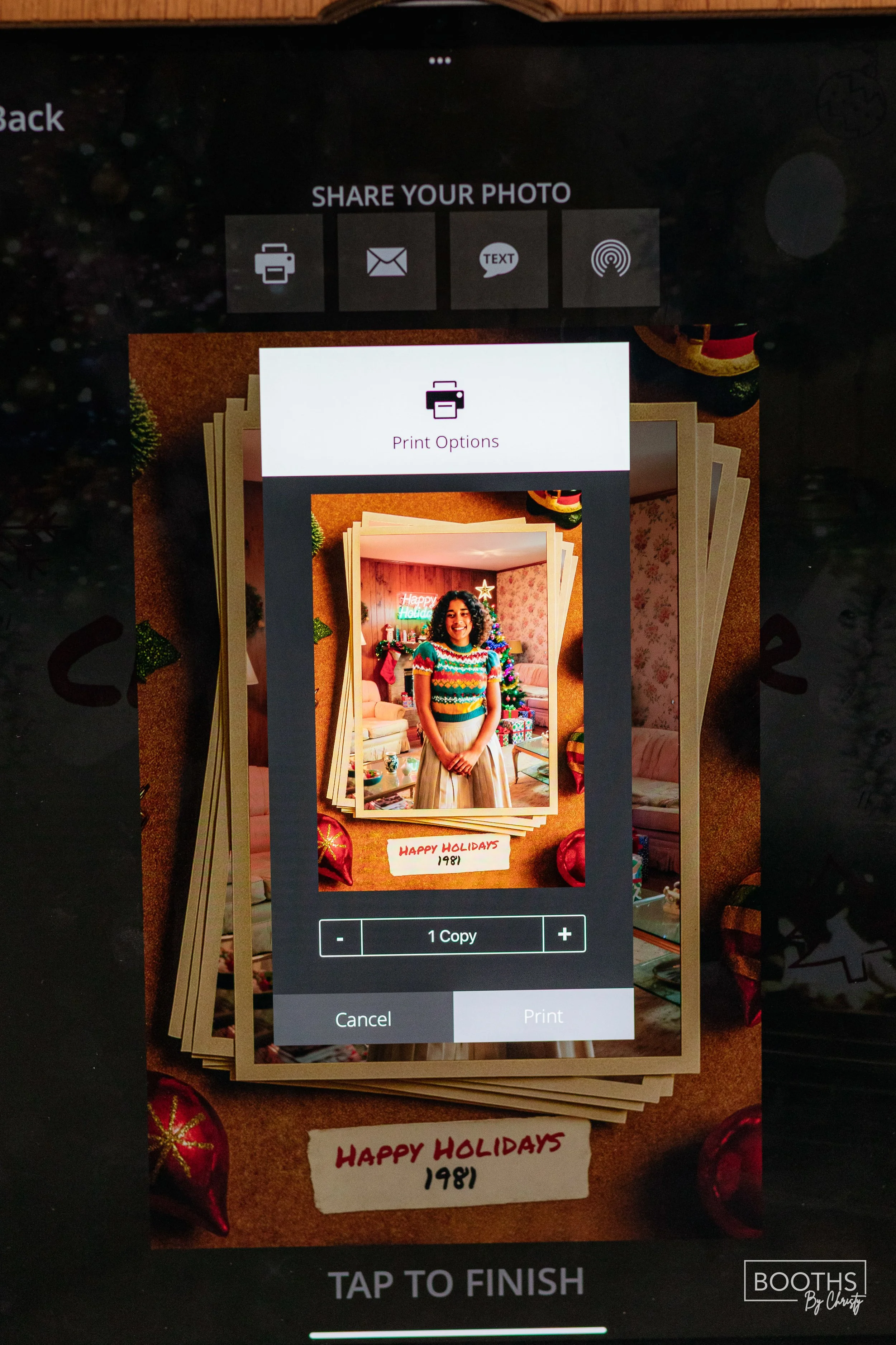 A digital screen showing a photo editing and printing app with a Christmas photo of a woman smiling, standing in a decorated room with a Christmas tree, presents, and holiday decorations in the background. The photo has a handwritten note underneath