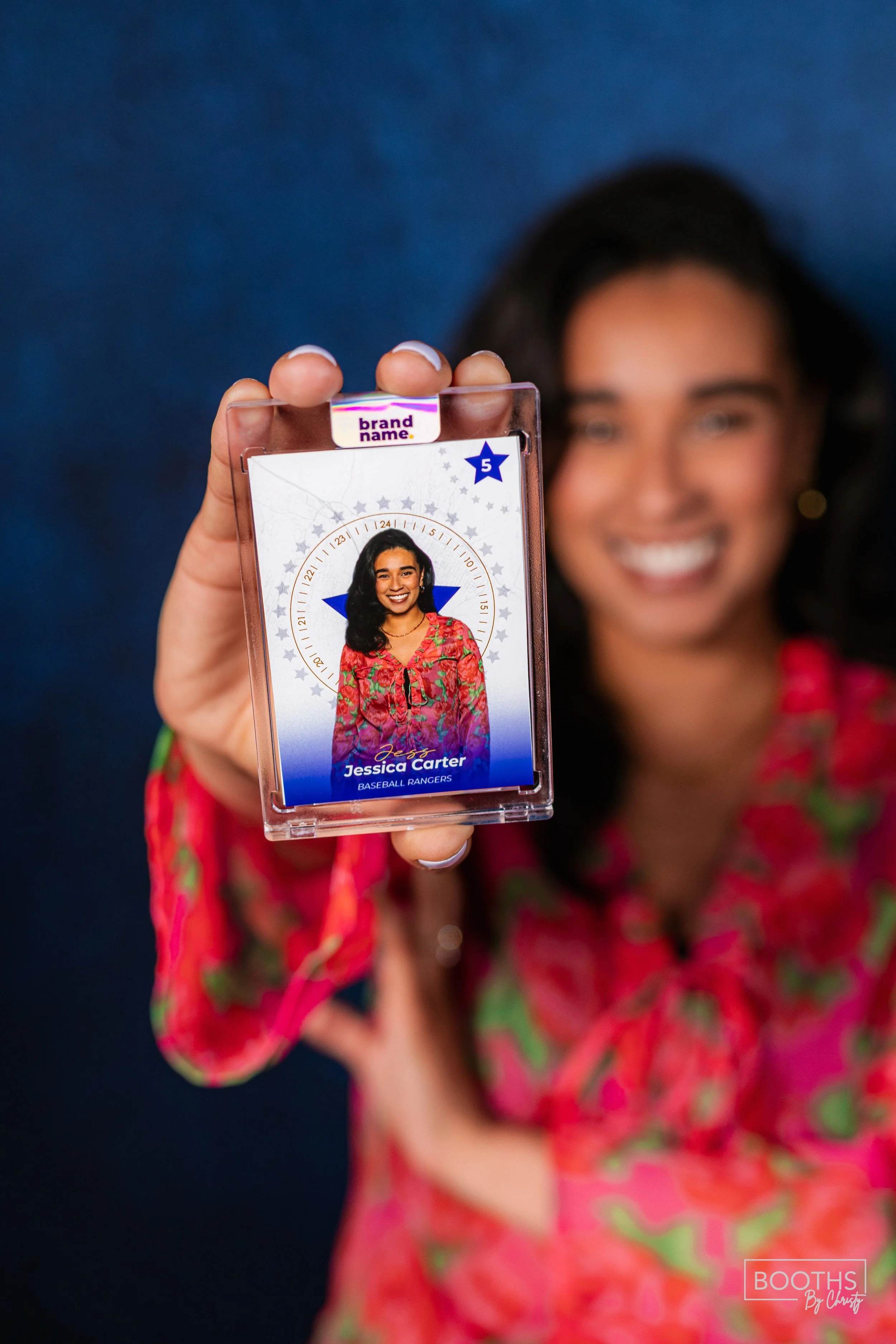 A woman with dark hair and a floral blouse holds up a clear plastic badge holder with a sports ID. The ID displays her picture, name 'Jessica Carter', and her team 'Baseball Rancers'. She has a big smile and the background is blurred, emphasizing the