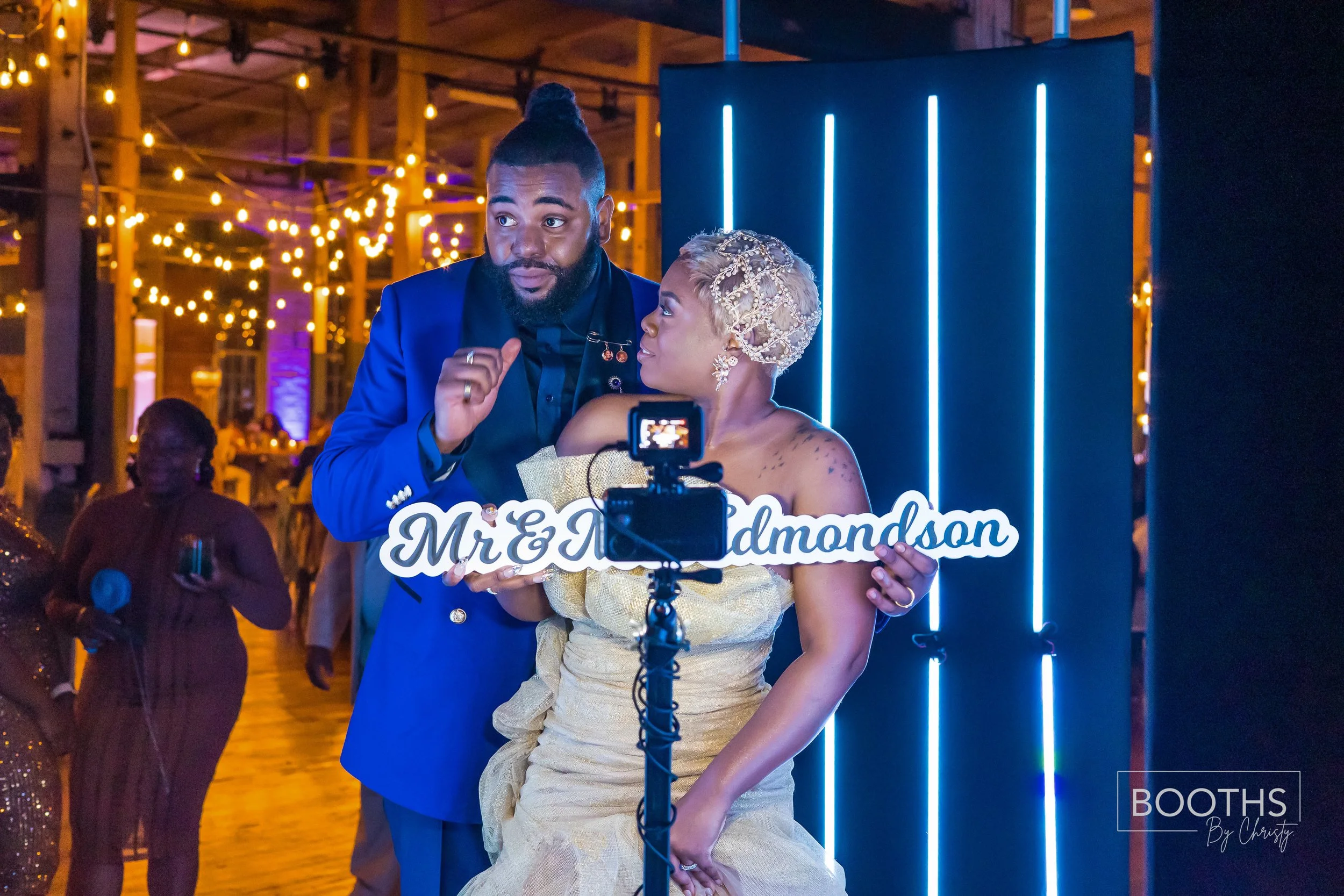 A couple at a wedding reception holding a sign that reads 'Mr & Mrs Diamondson'; the woman is wearing a gold dress and a decorative headpiece, the man is in a blue suit. They are standing in front of a blue illuminated backdrop with warm, string ligh