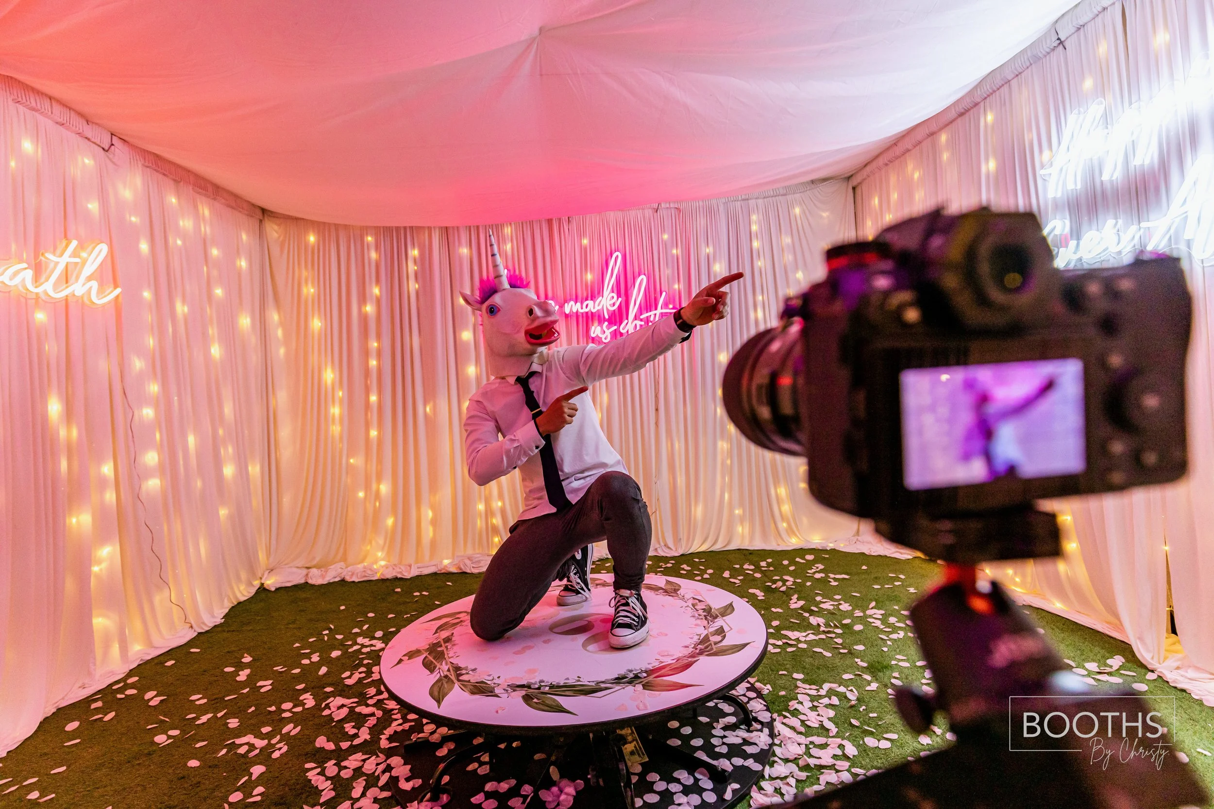 A person wearing a unicorn mask and casual clothes, kneeling on a circular platform, pointing and posing for a camera inside a decorated party booth with pink and white drapes, string lights, and neon signs.
