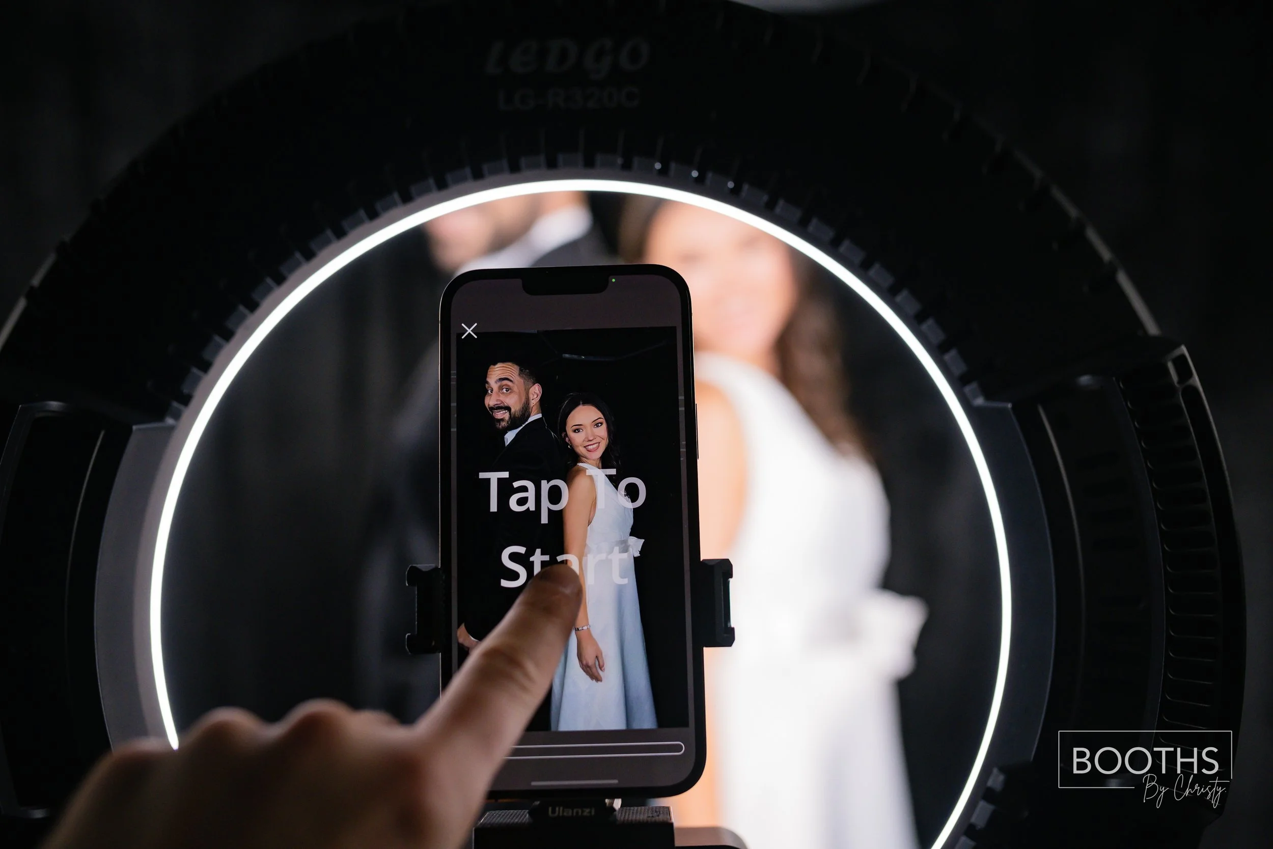 A person holding a smartphone with on-screen text that says "Tap To Start" in front of a wedding photo of a smiling bride and groom. The photo is framed by a circular LED ring light.