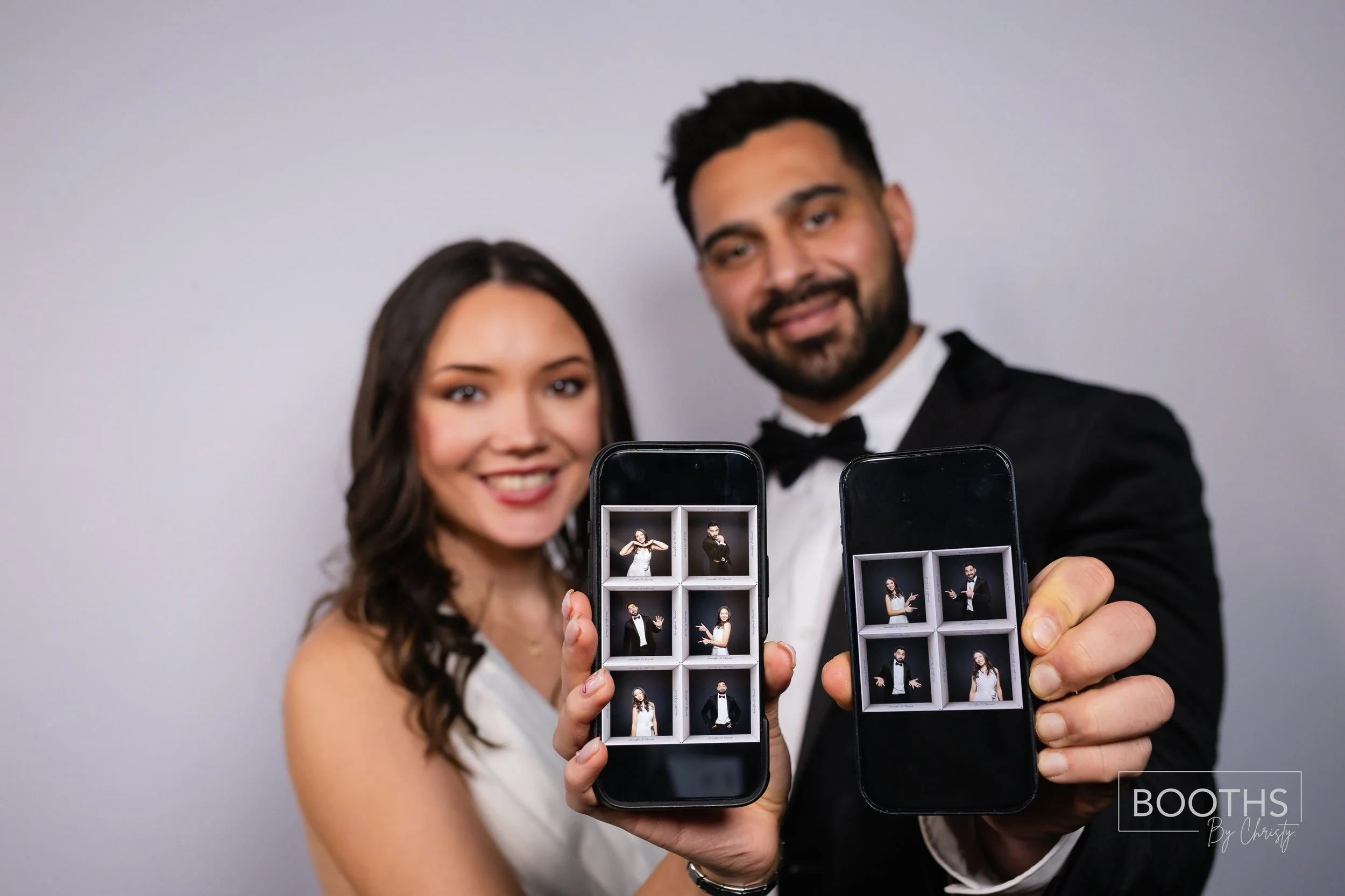 A couple in formal wear taking selfies with their phones, showing photo booth pictures of themselves wearing tuxedo and dress, on a plain gray background.