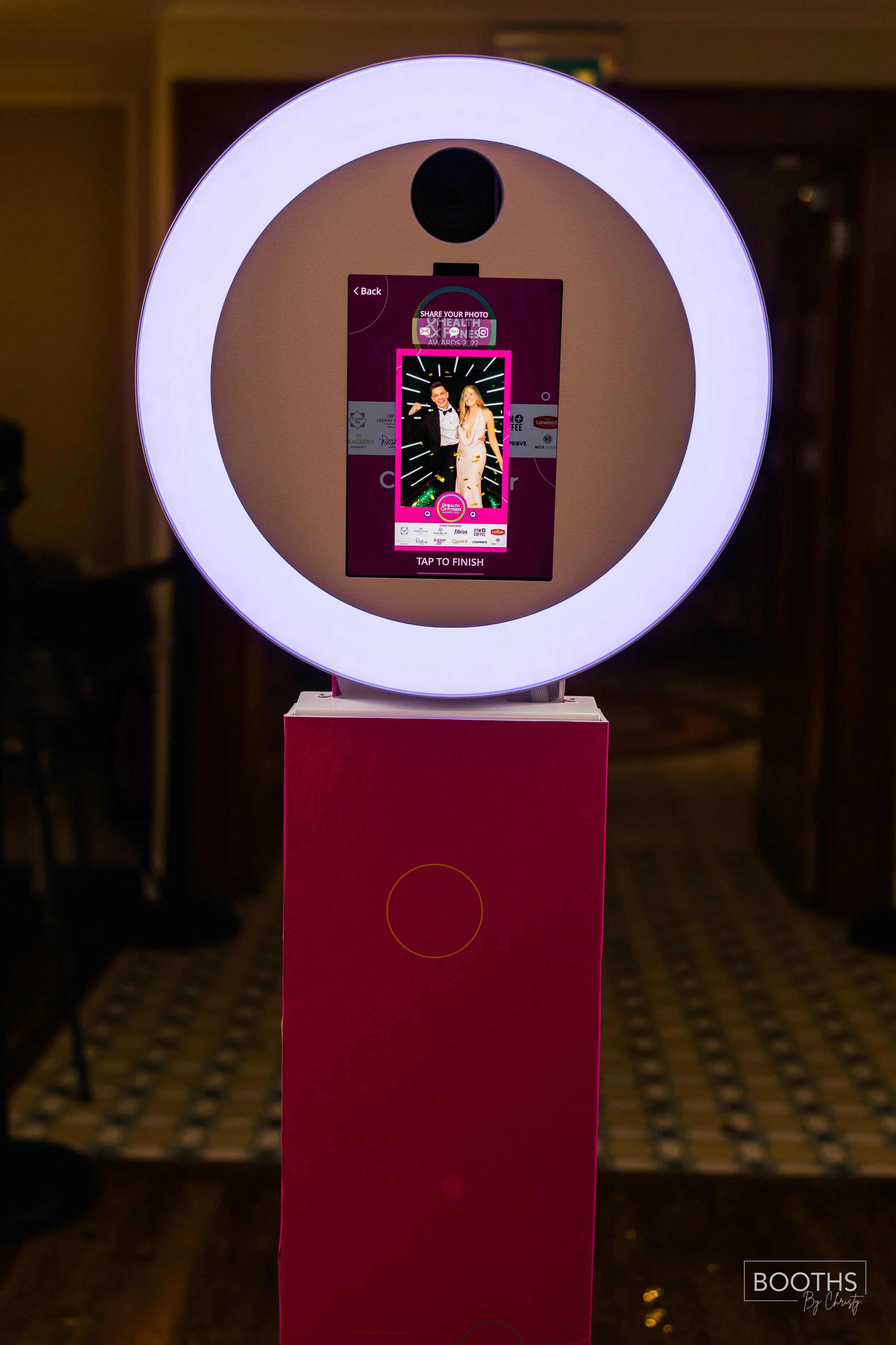 Photo of a photo booth setup with a circular ring light surrounding a screen displaying a photo of a man in a tuxedo and a woman in a white dress at an event called Share Your Photo and Health Expo.
