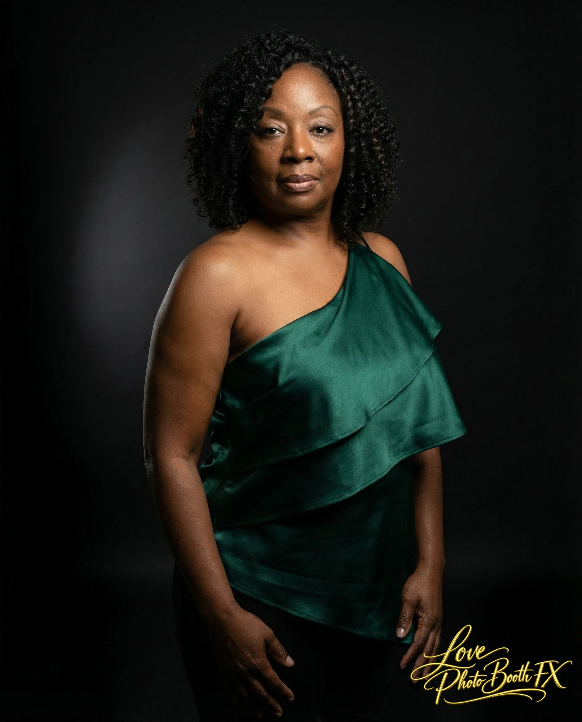A woman with dark skin and curly black hair poses against a black background. She is wearing a green satin one-shoulder top and has a serious expression. The photo has a professional studio lighting and is marked with 'Love Photo Booth FX' in cursive