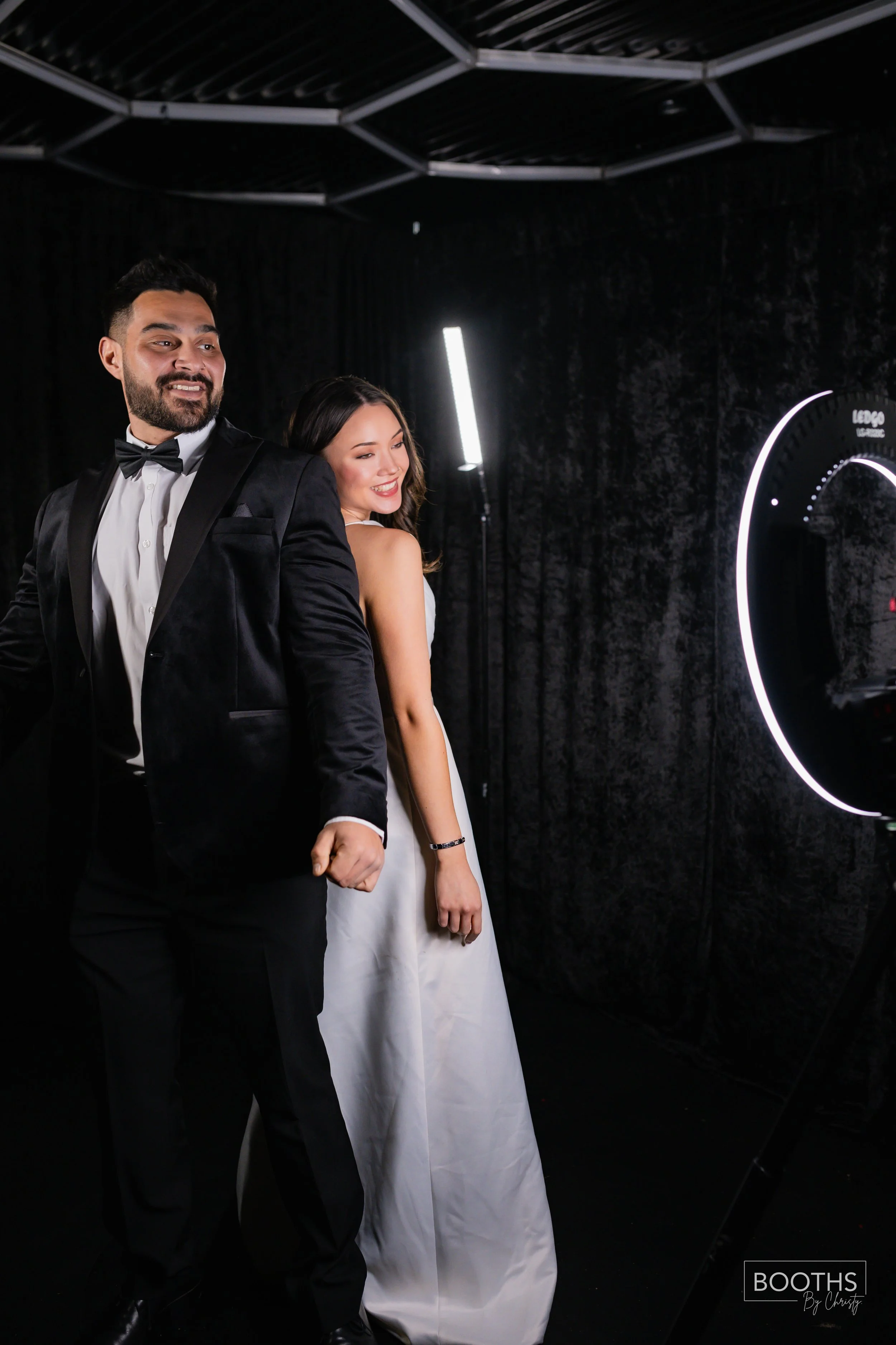 A man and woman dressed in formal attire posing in a photo booth with a black background and neon light ring.