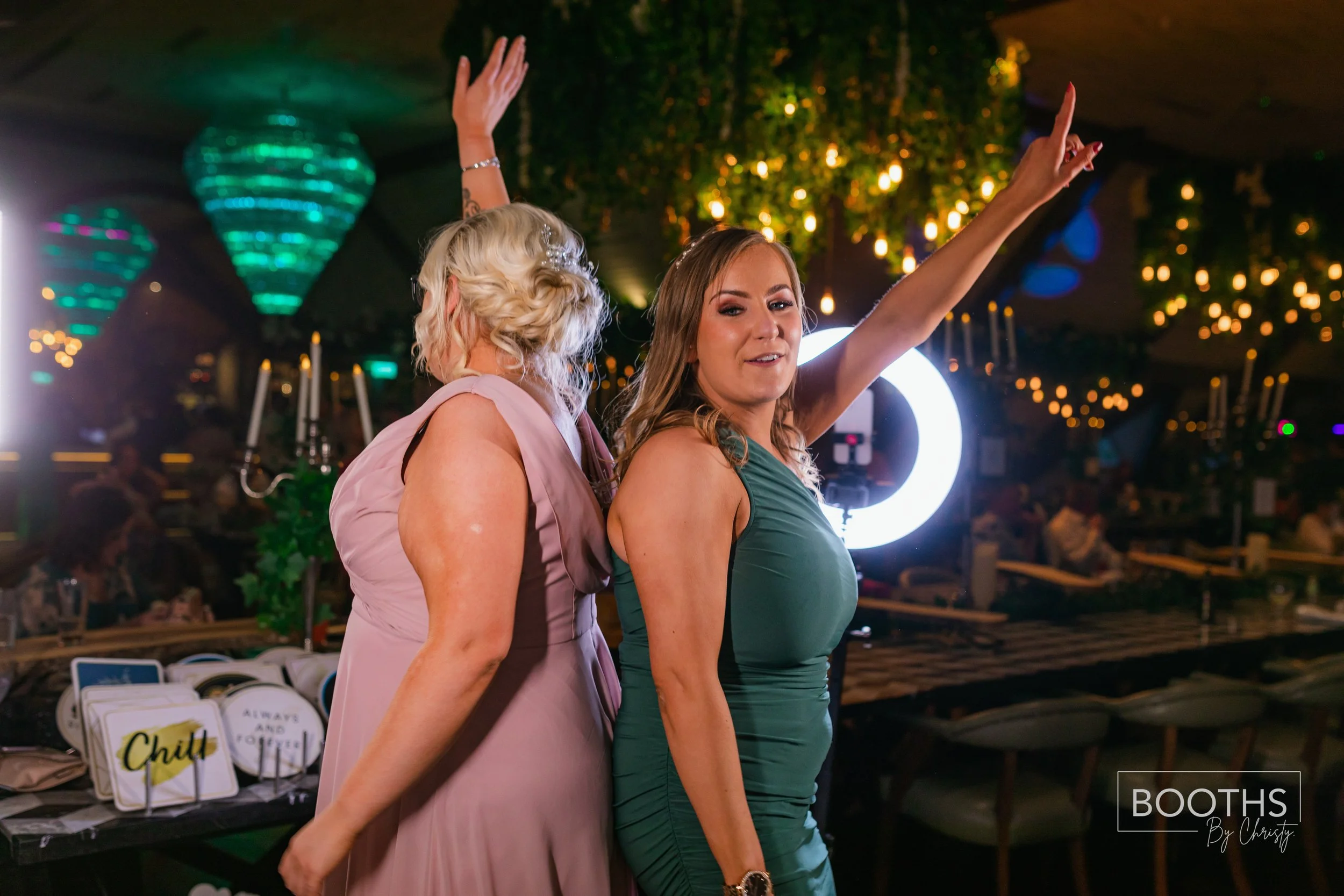 Two women dancing and having fun at a party, with one woman in a pink dress and the other in a green dress, under festive lighting and decorations.