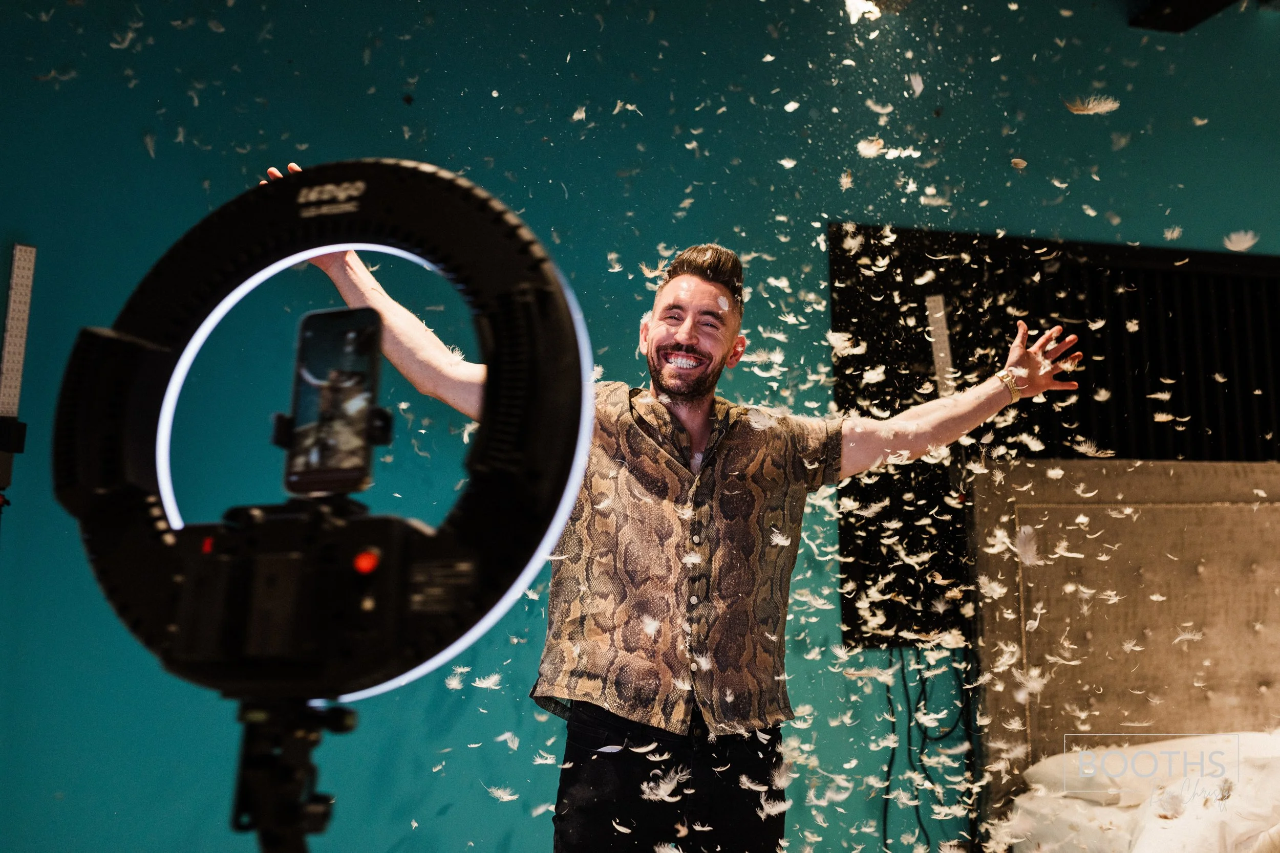 A man with a beard and short hair smiling and spreading his arms at a celebration with feathers flying around, as he records the scene using a smartphone mounted on a ring light with a camera. The background includes a teal wall, a large speaker, and