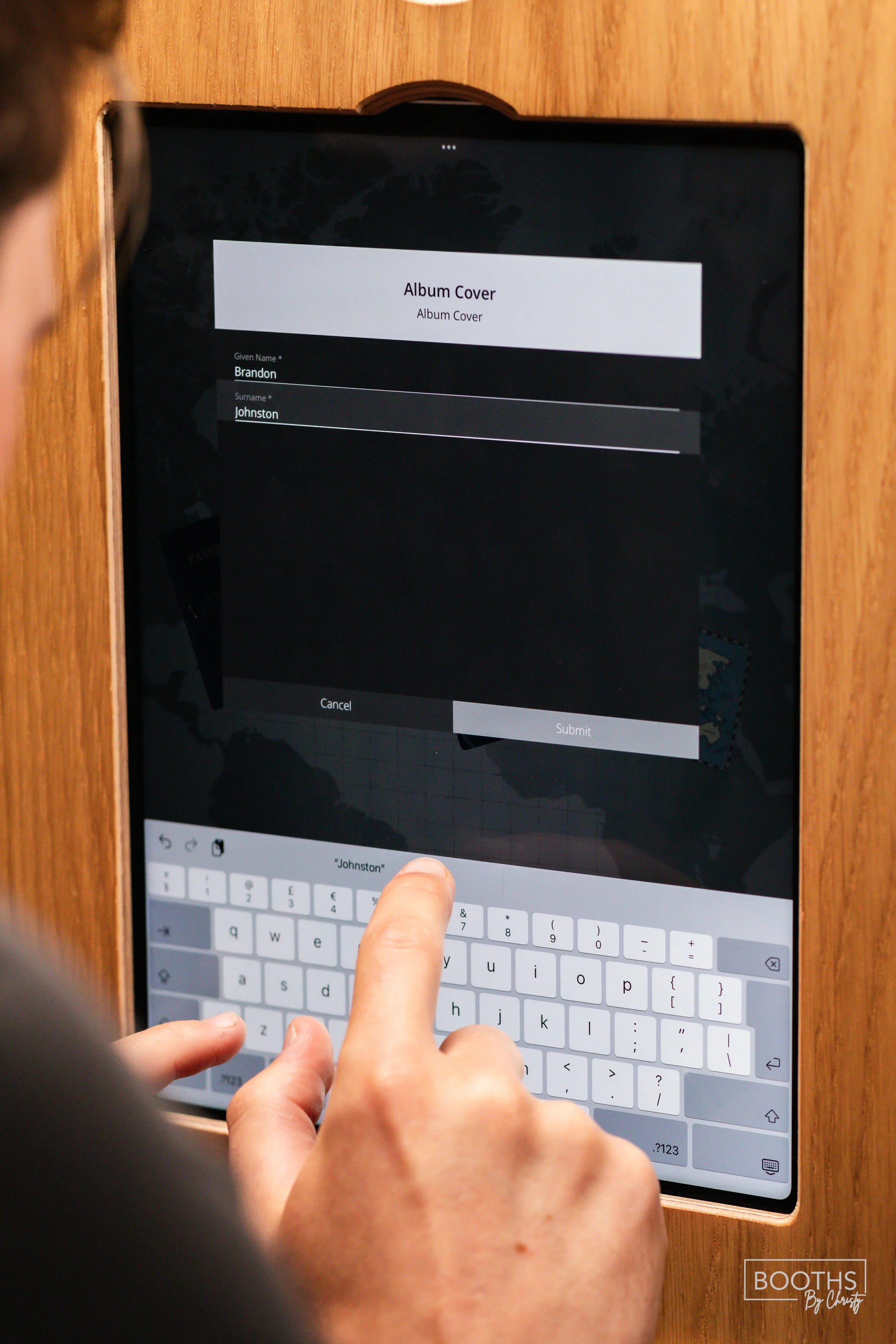 A person entering the name 'Brandon Johnston' on a tablet screen in a wooden frame, labeled 'Album Cover'.