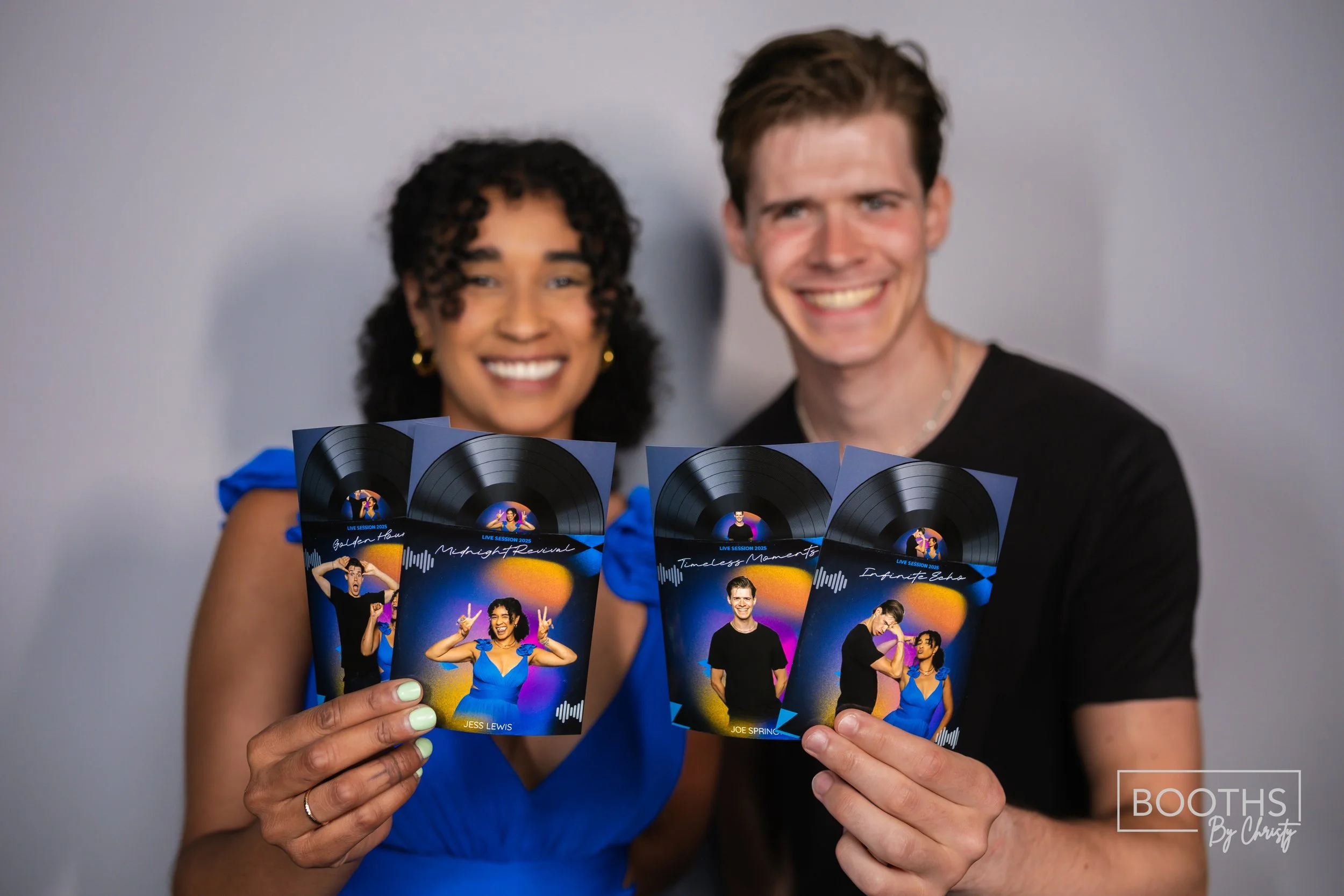 Two smiling people holding colorful event flyers, with a gray background. The woman is wearing a blue dress and has curly black hair, and the man has short brown hair and is wearing a black T-shirt.