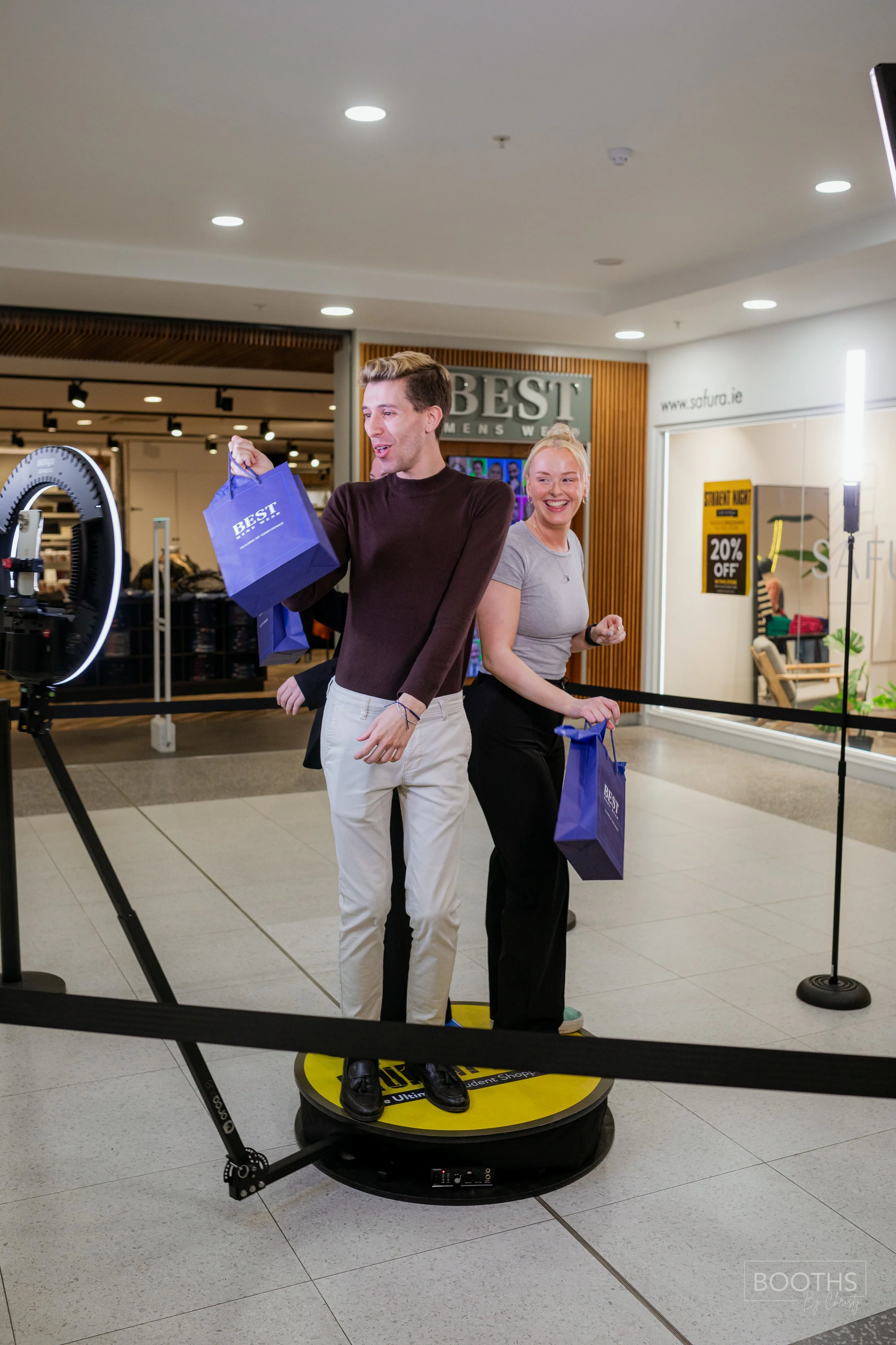 Two people standing on a yellow platform, smiling and holding shopping bags, during a promotional event in a shopping mall.