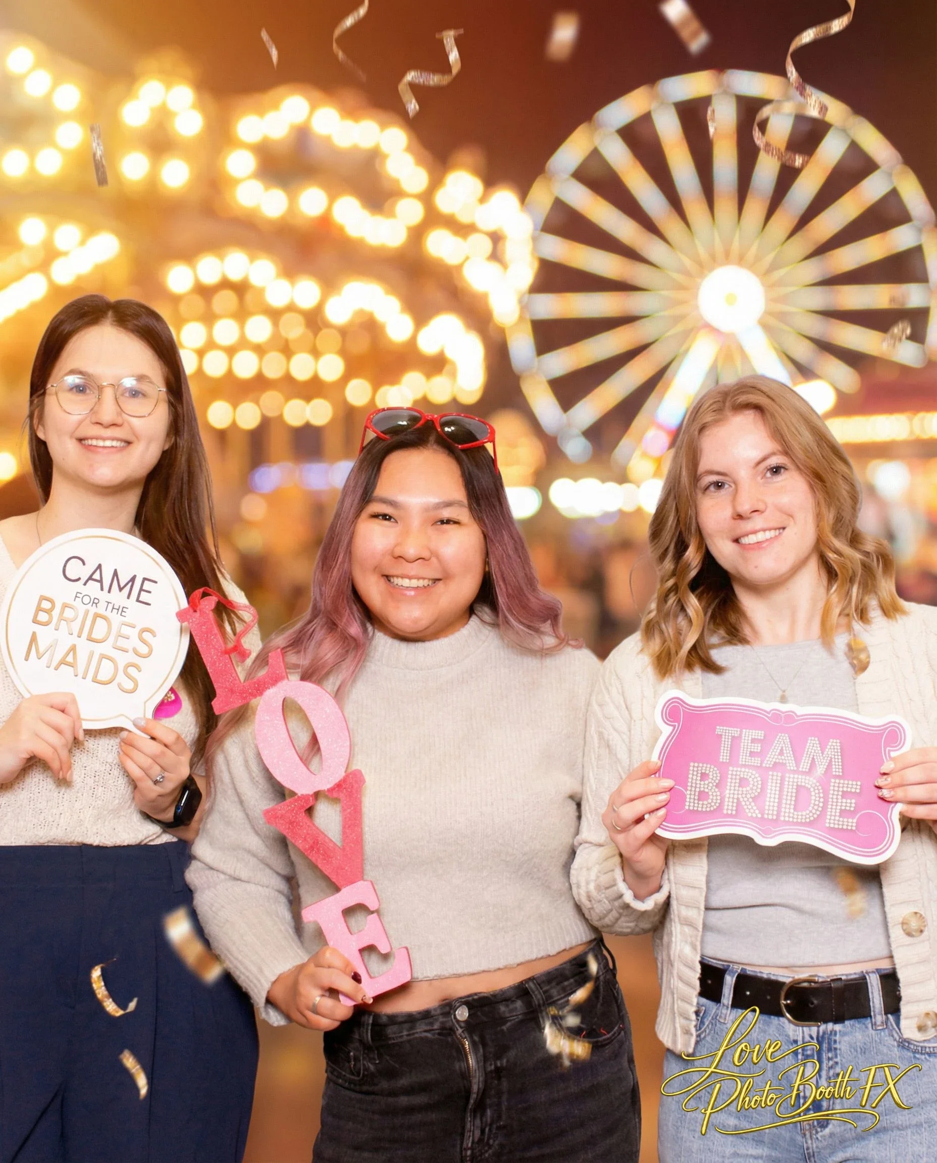 Three women at a fair or carnival at night, holding signs supporting bridesmaids and the bride. The woman on the left holds a sign that says "CAME FOR THE BRIDES MAIDS," the woman in the center holds pink and white letters spelling "LOVE," and the wo