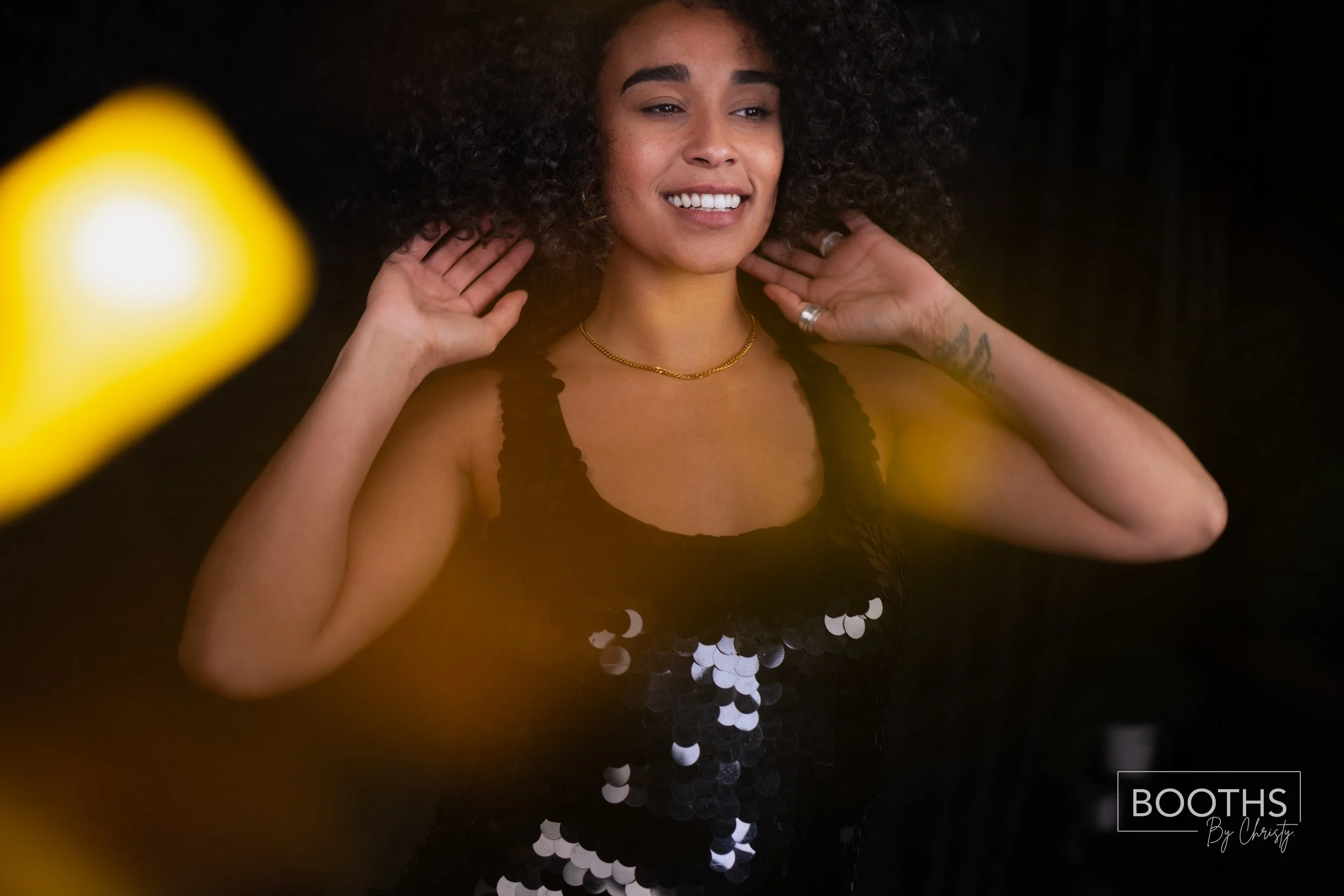 A woman with curly hair smiling and adjusting her earrings, wearing a black sequined top, gold necklace, and rings. The photo is taken against a dark background with some yellow blurred objects in the foreground.