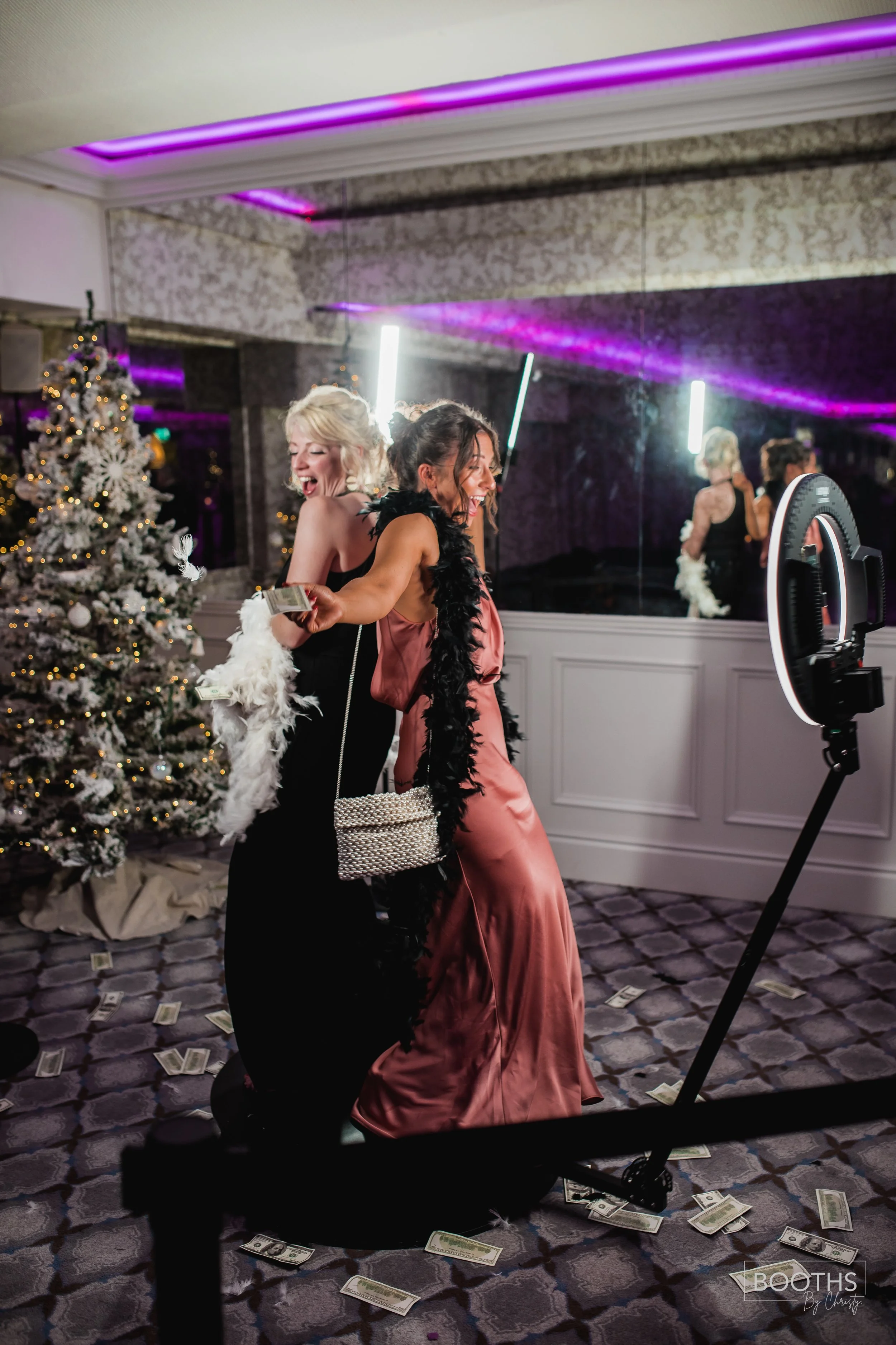 Two women dancing and laughing in a decorated room with a Christmas tree, surrounded by dollar bills on the floor, with a ring light and mirror in the background.