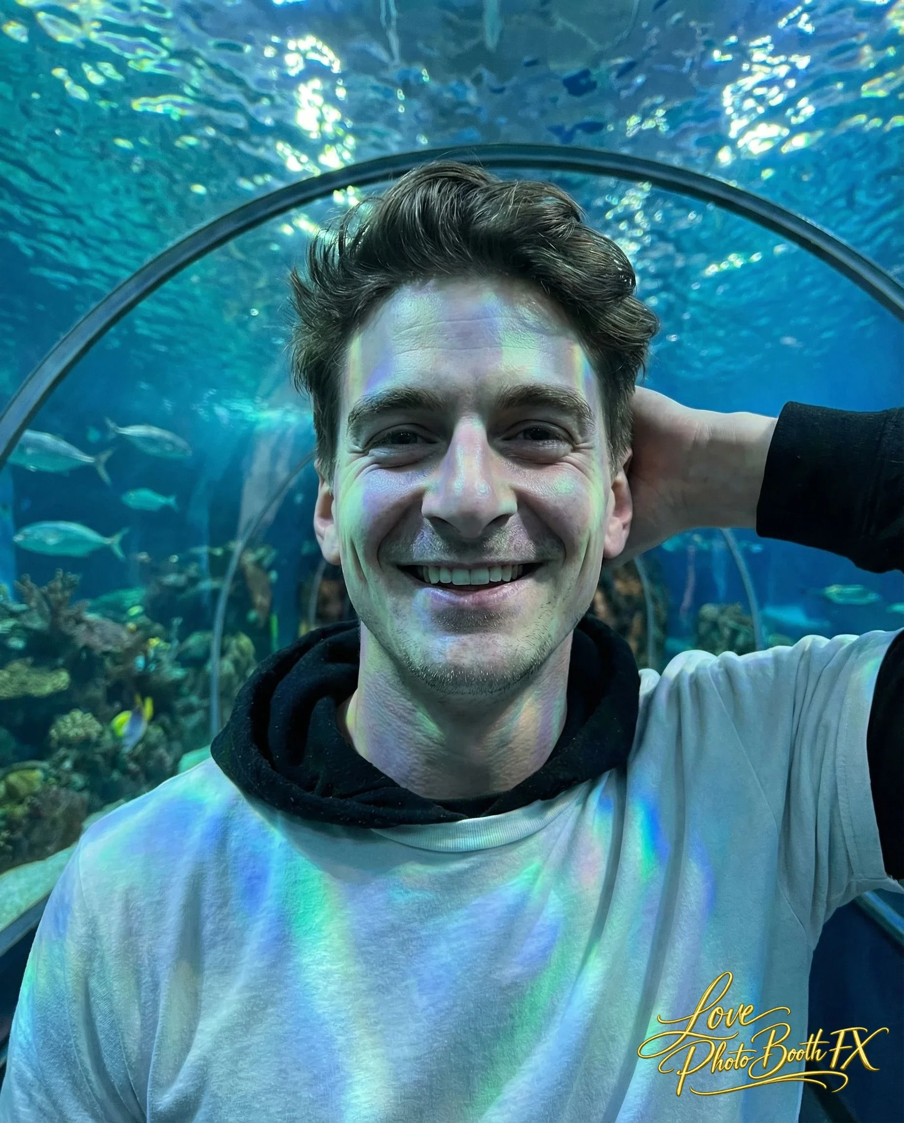 A young man smiling at the camera inside an aquarium tunnel with fish swimming around.
