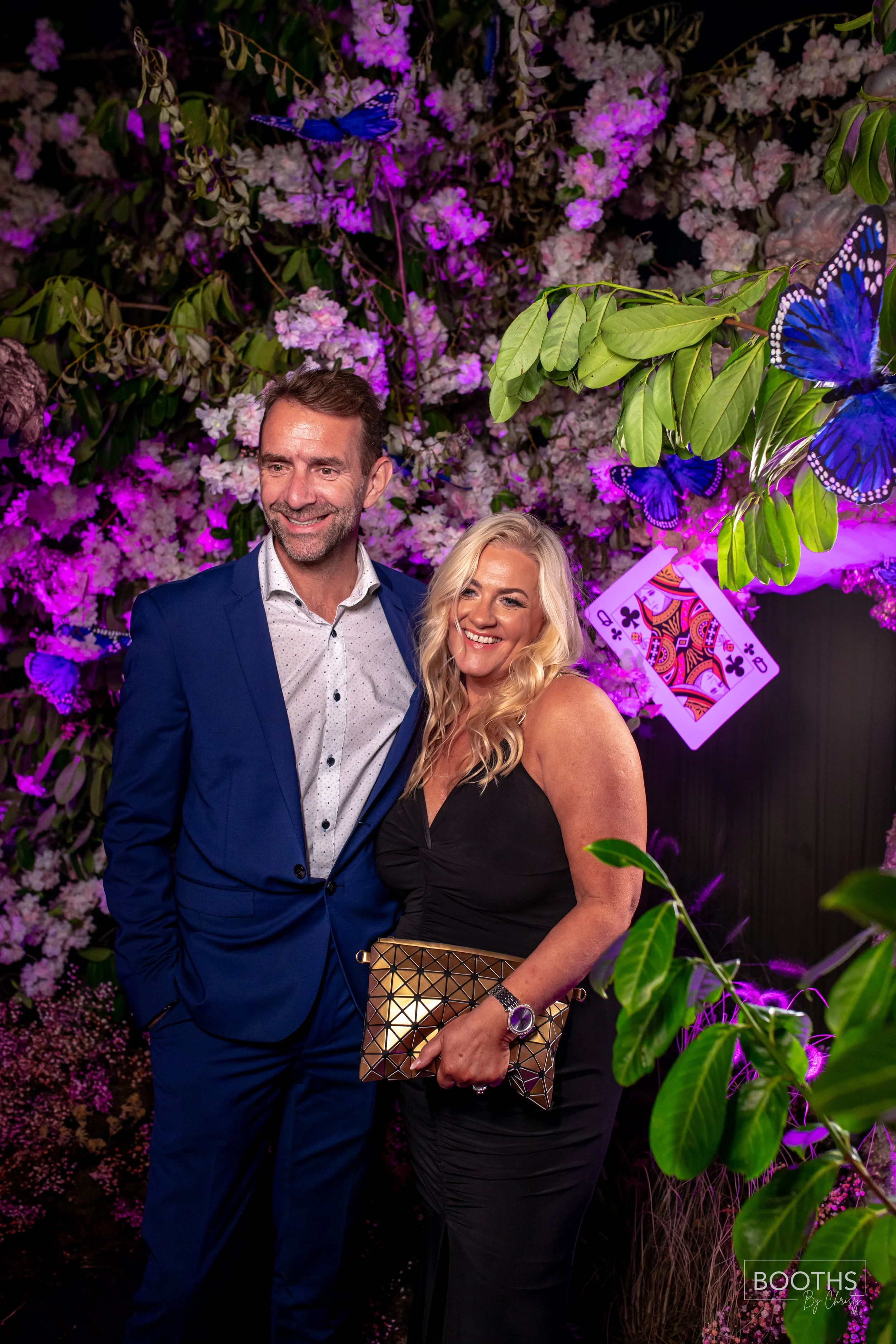 A smiling man in a blue suit and woman in a black dress posing together at an event with purple lighting, surrounded by greenery, flowers, butterfly decorations, and playing card decorations in the background.