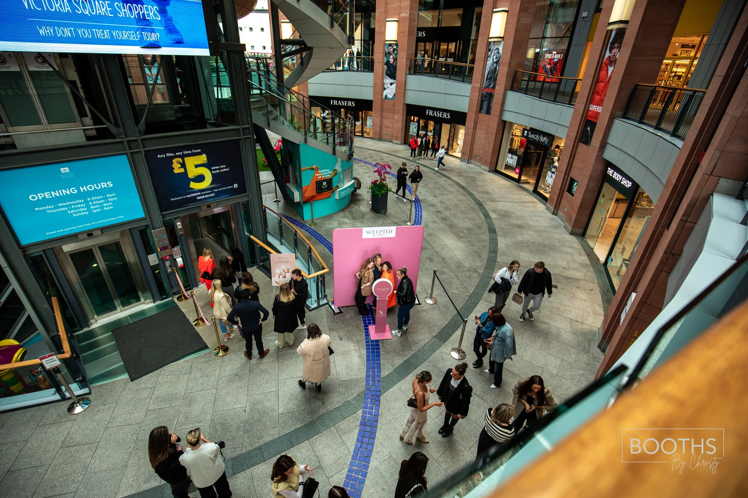 Interior view of a shopping mall with multiple floors, shops, and people walking or gathering in groups. There are digital screens displaying store opening hours and advertisements, and a pink display stand in the center with a group of people taking