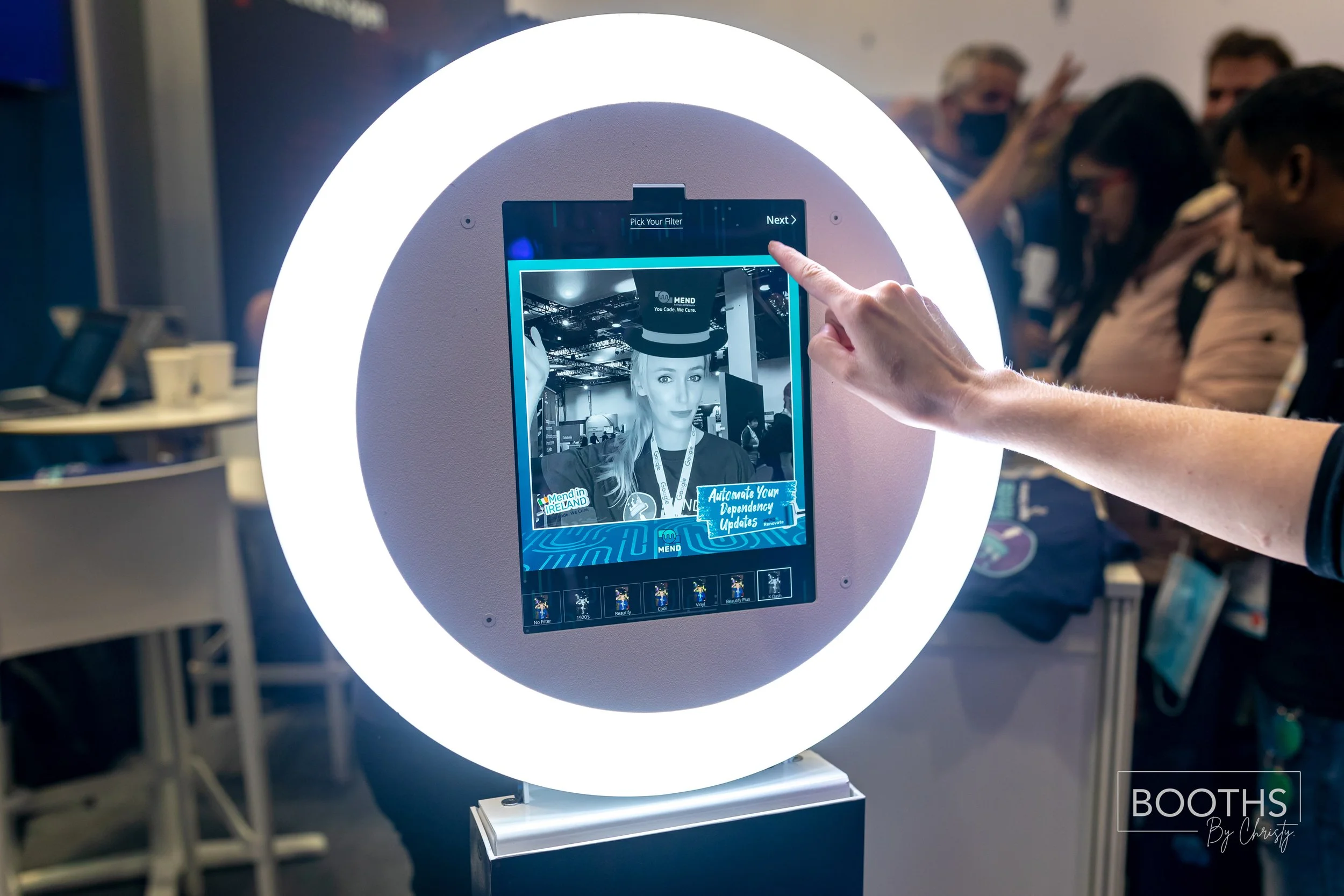 A person interacting with a digital photo booth screen surrounded by a brightly lit ring, displaying a monochrome portrait of a woman wearing a hat at an event.