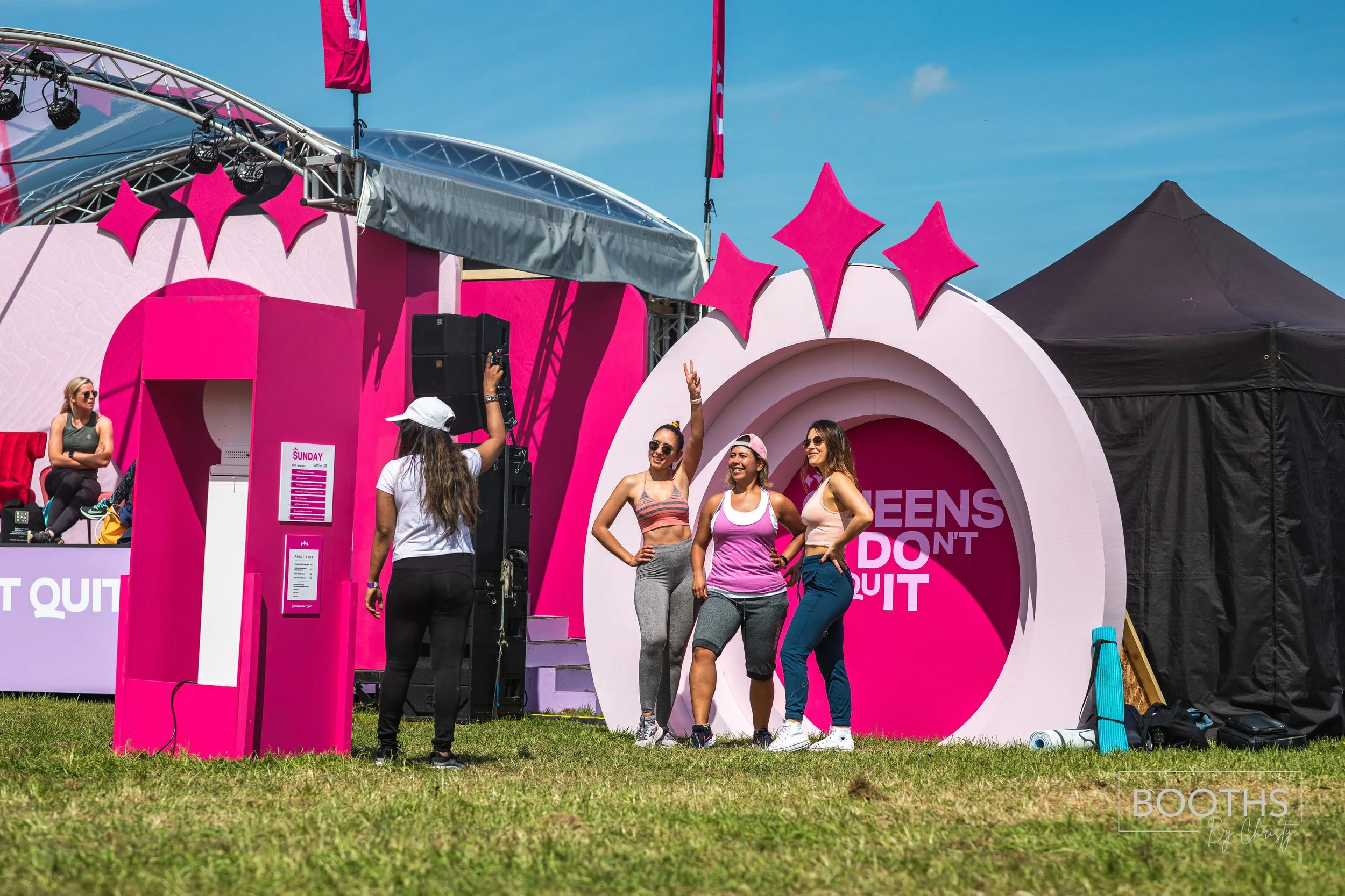 Group of women posing in front of a pink and white event booth with a sign that reads 'Queens Don't Quit' at an outdoor festival on a sunny day.