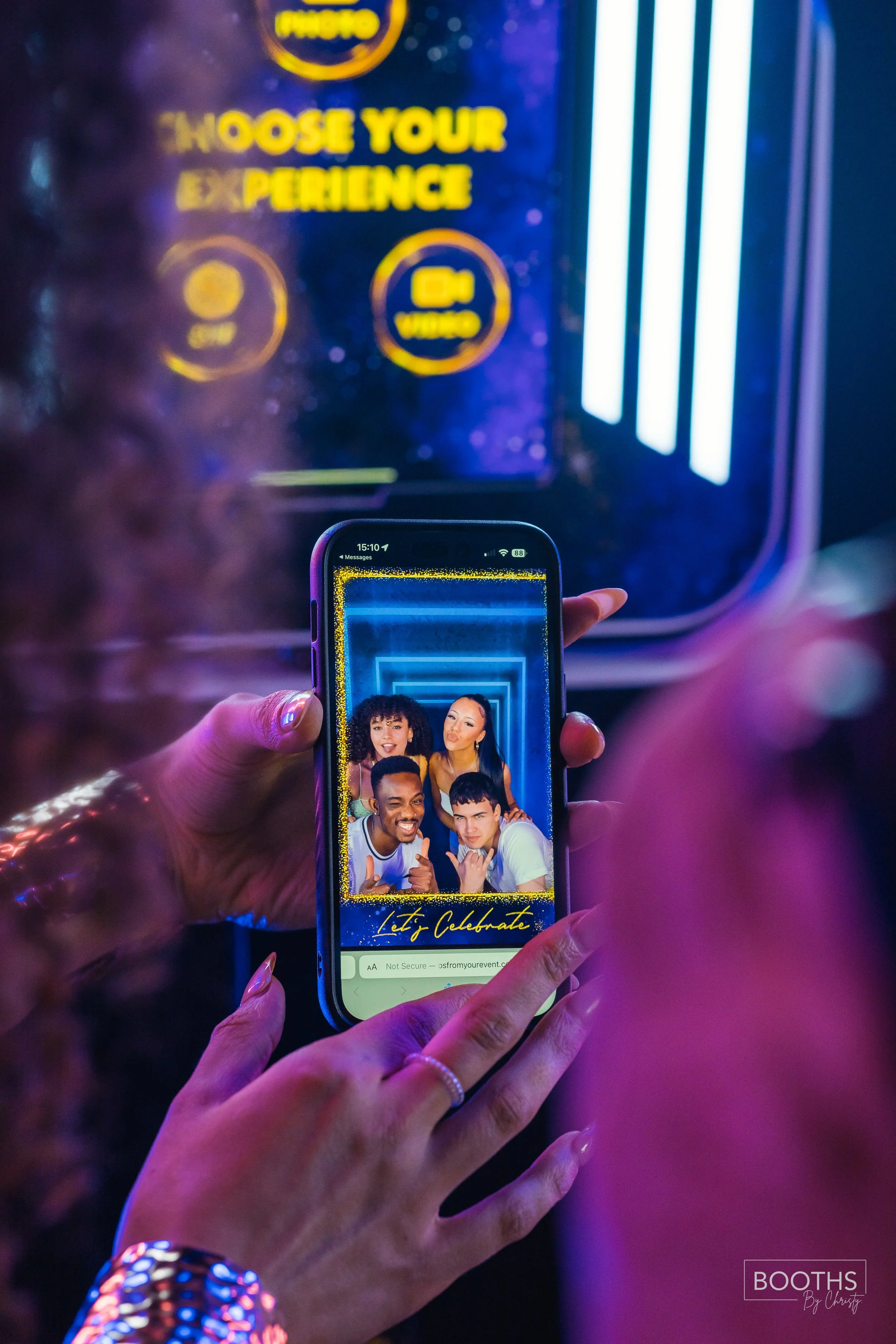 Person holding a smartphone taking a group selfie with four smiling friends inside a photo booth, with creative blue neon lighting and a decorative frame that says "Let's Celebrate".