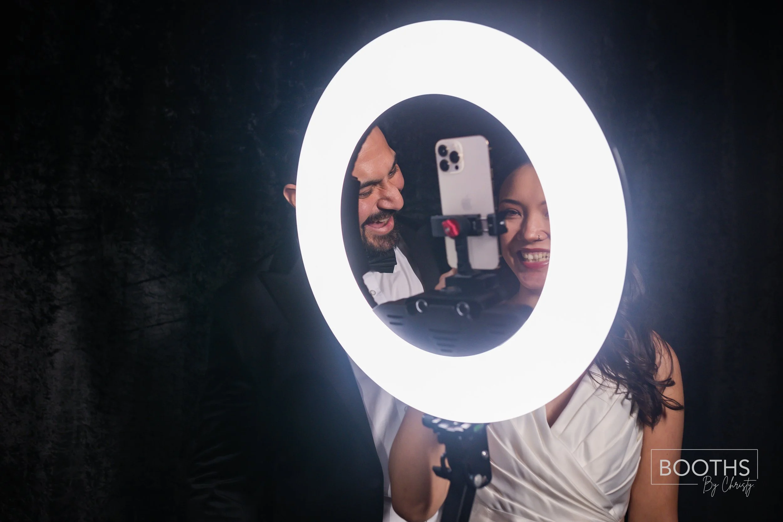 A man and woman looking at a smartphone mounted on a tripod within a bright ring light, smiling and laughing, with a black background.