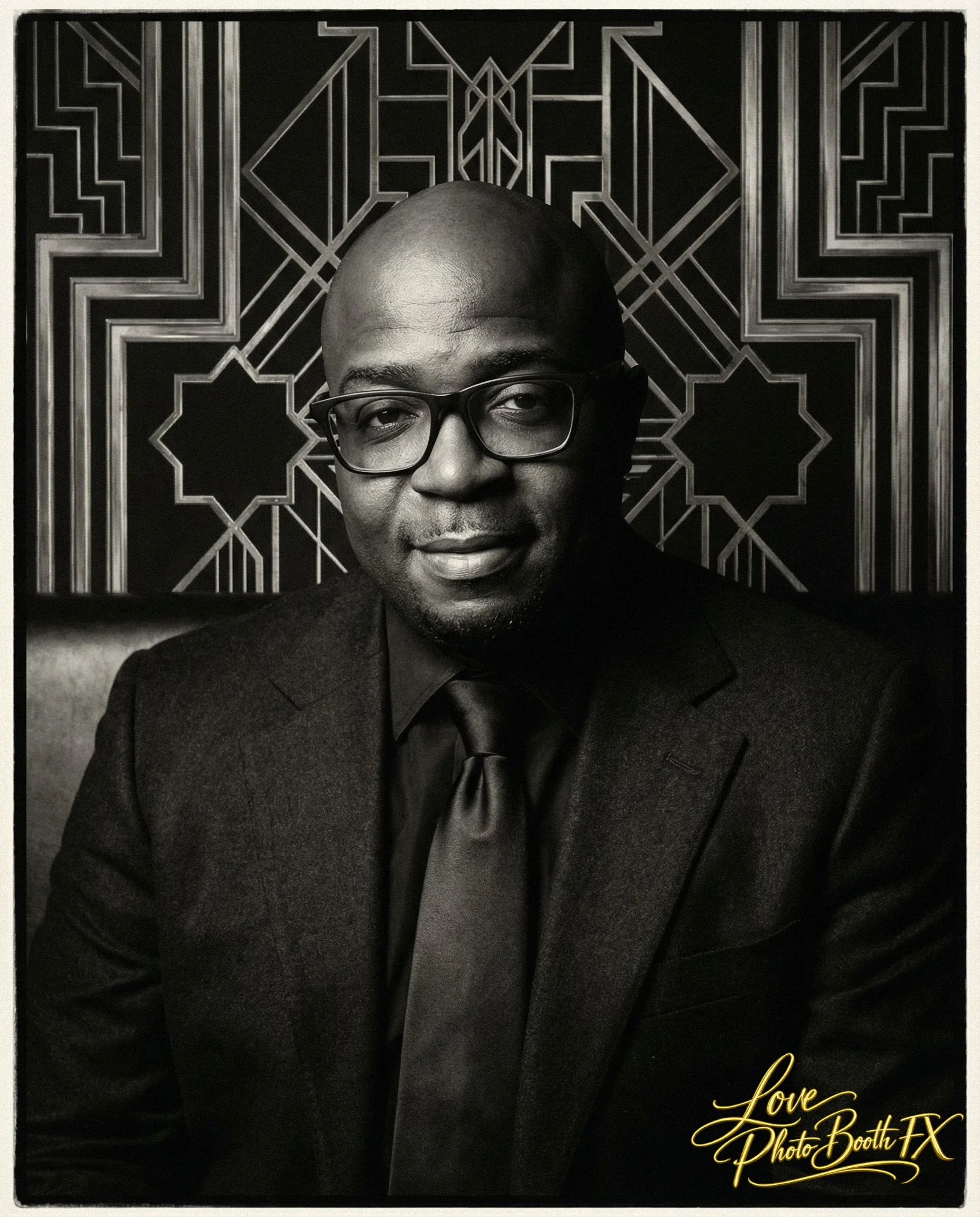 A black-and-white portrait of a man wearing glasses and a dark suit with a tie, seated in front of an ornate, geometric patterned background.