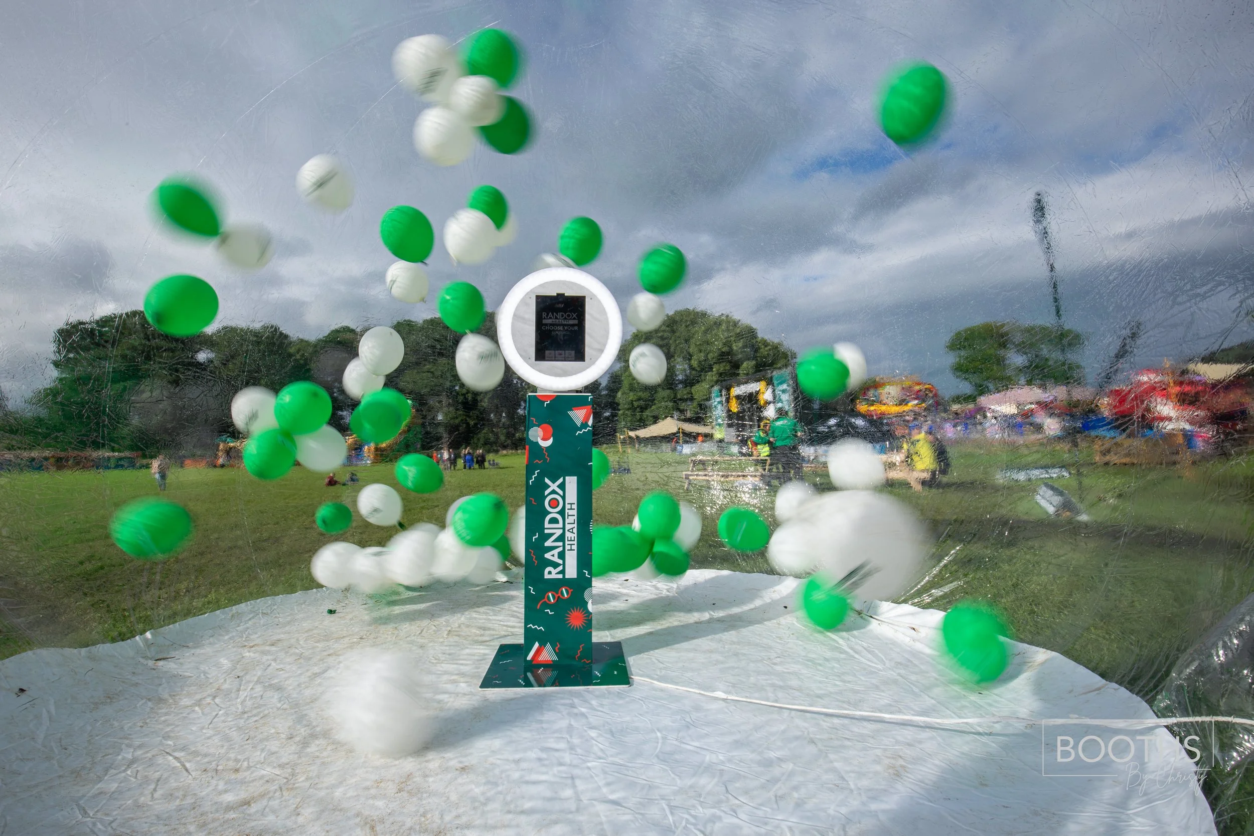 View through a transparent surface showing green and white balloons being released into the air at an outdoor event, with trees, tents, and people in the background.