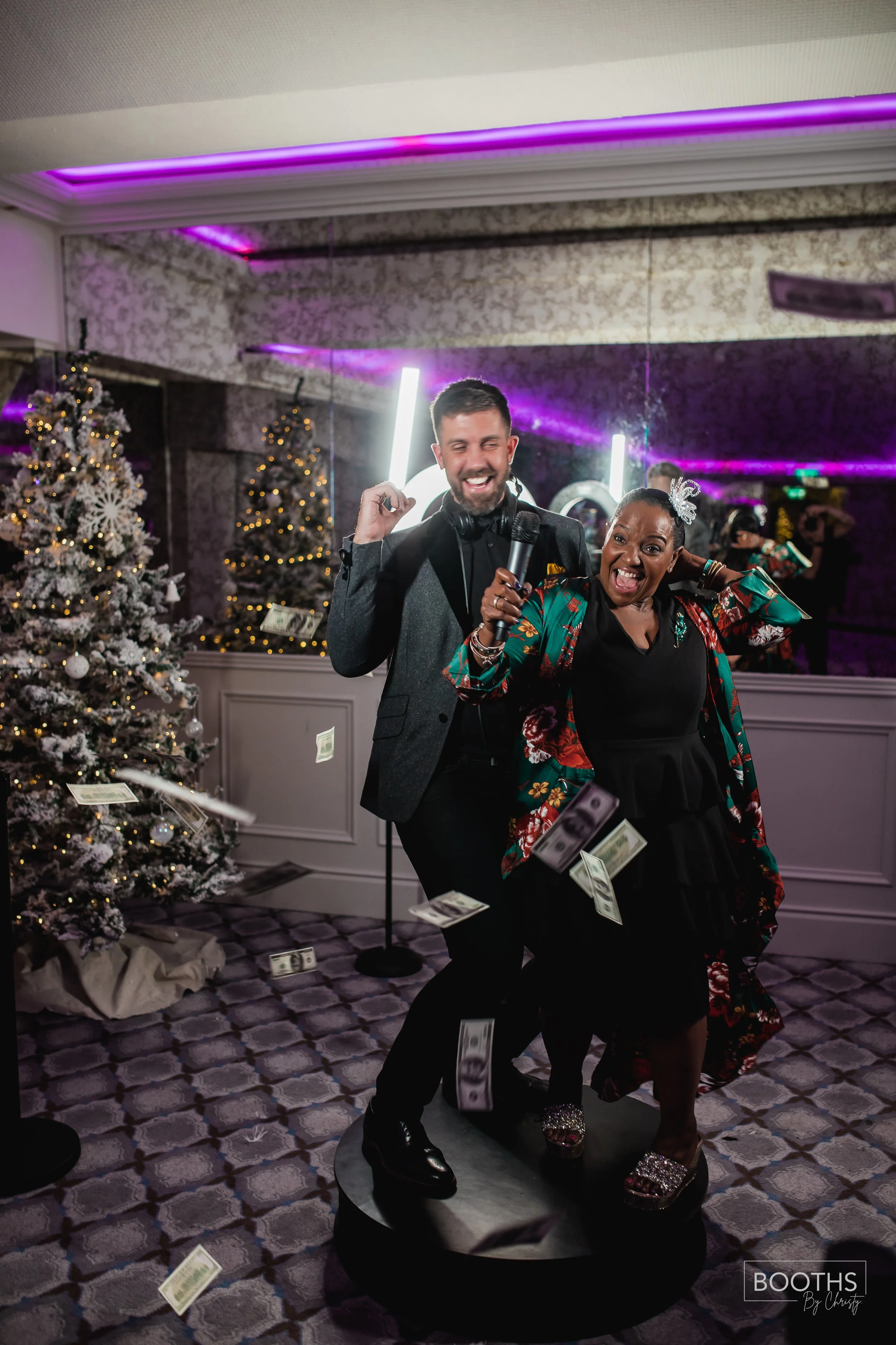 Two people dancing and having fun at a festive party with Christmas trees in the background, falling dollar bills, and colorful lighting.