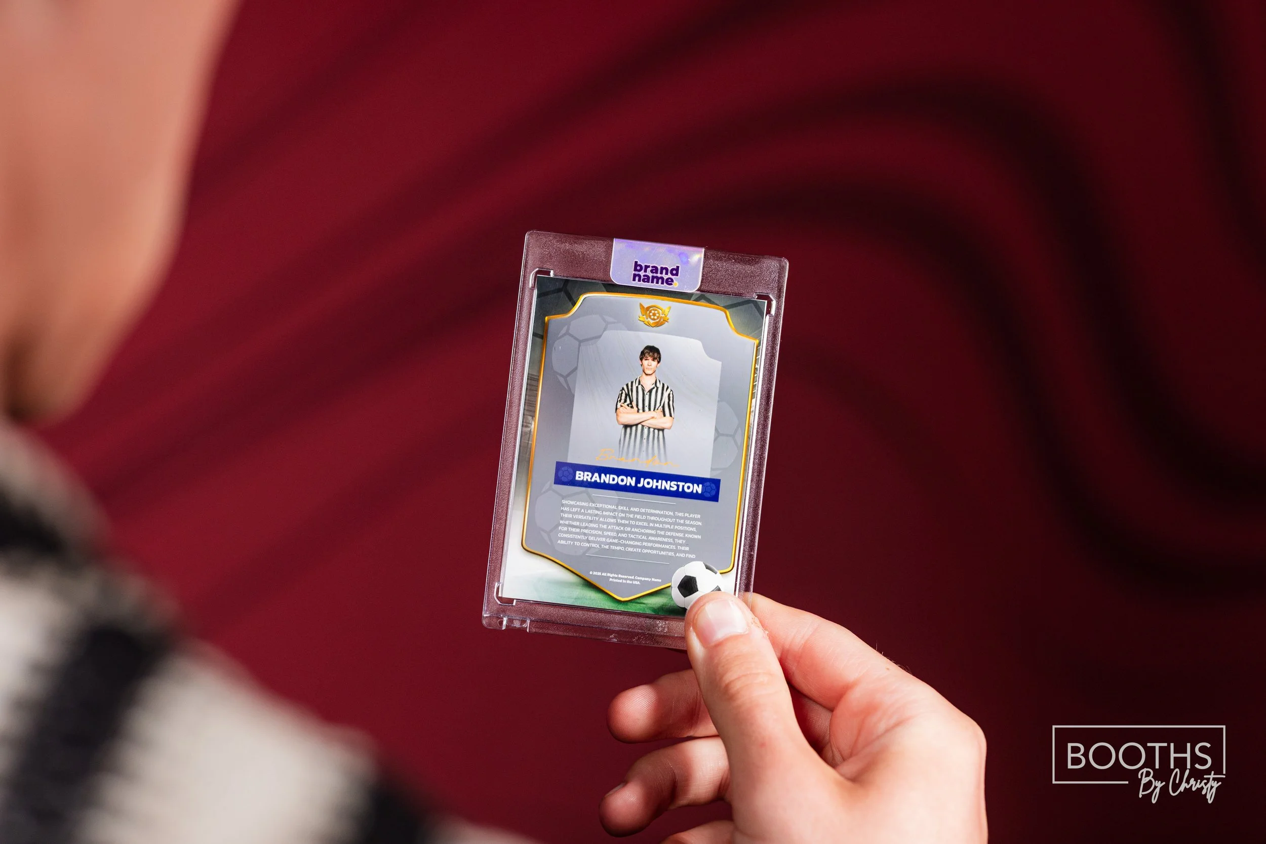 A close-up of a person holding a player card with a photo of a man in a striped shirt, named Brandon Johnston, inside a transparent plastic case with a small soccer ball on the bottom. The background is blurred with red tones.