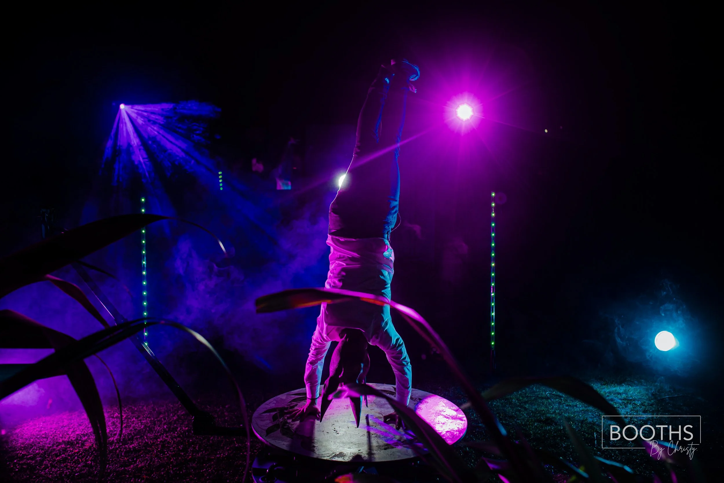 A person performing a handstand on a circular platform at a nightclub or party with vibrant purple, pink, and blue lights and smoke effects.