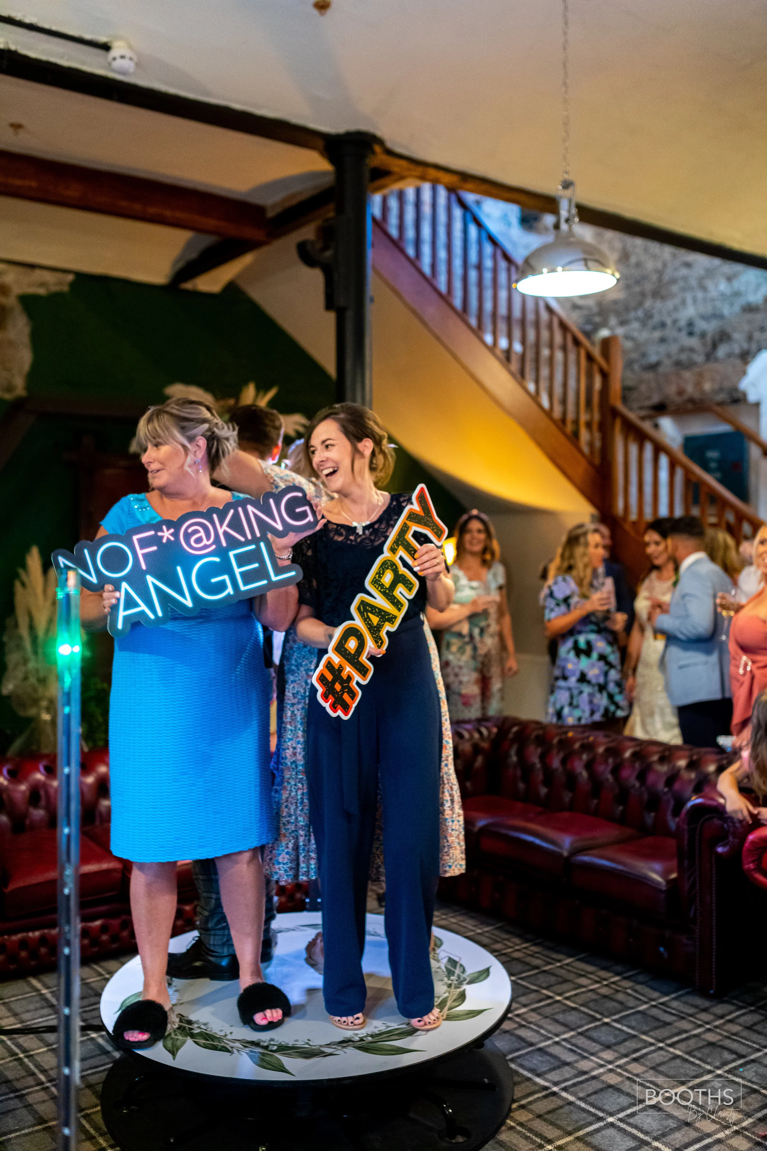 Two women at a celebration holding neon signs that read 'NO F*KING ANGEL' and '#PARTY,' standing on a circular platform in a room with guests in the background.