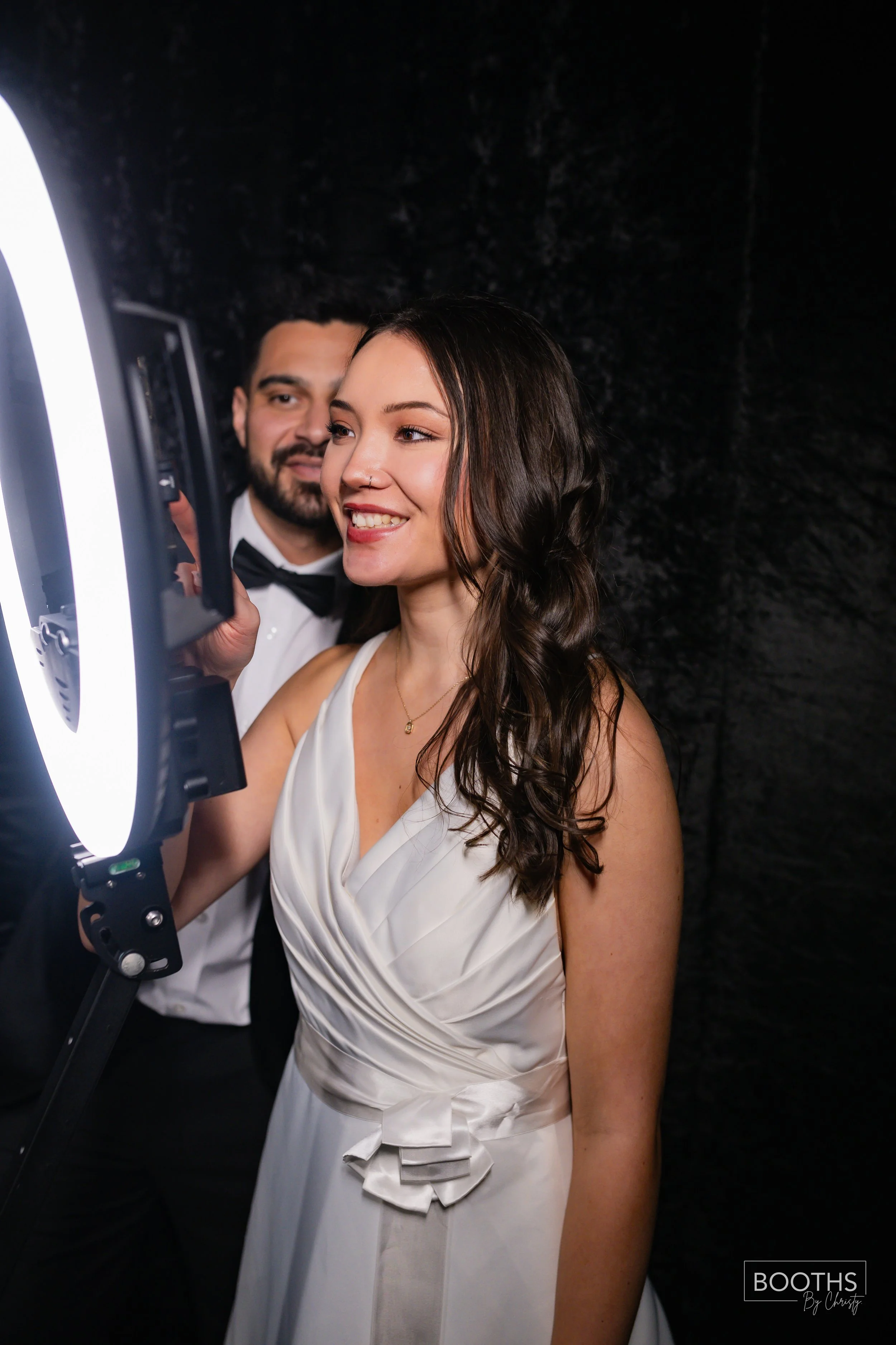 A woman in a white dress and a man in a tuxedo taking a photo together using a ring light. The woman is smiling and the man is in the background.