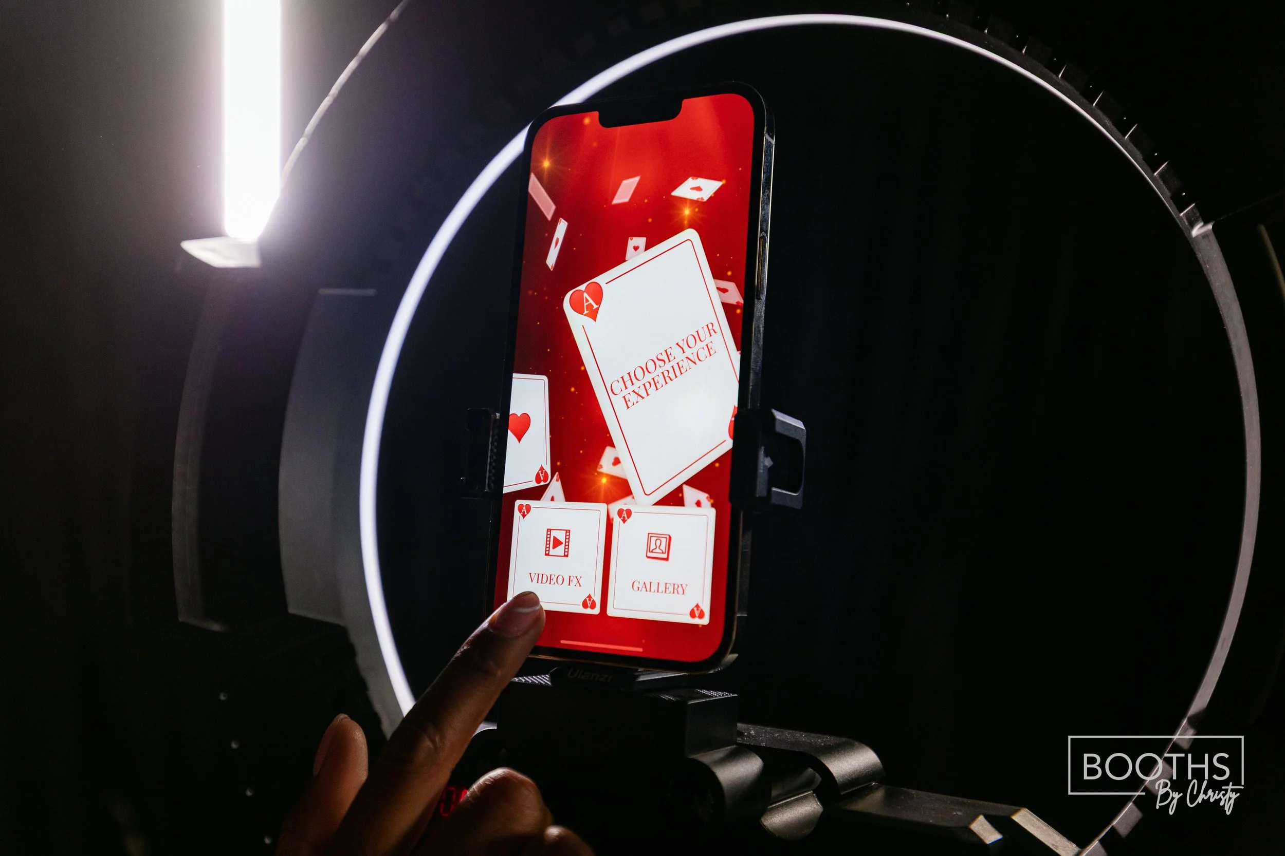 A smartphone displaying a poker game app with options for 'Choose Your Experience,' 'Video FX,' and 'Gallery,' mounted inside a circular light ring with a person touching the screen.
