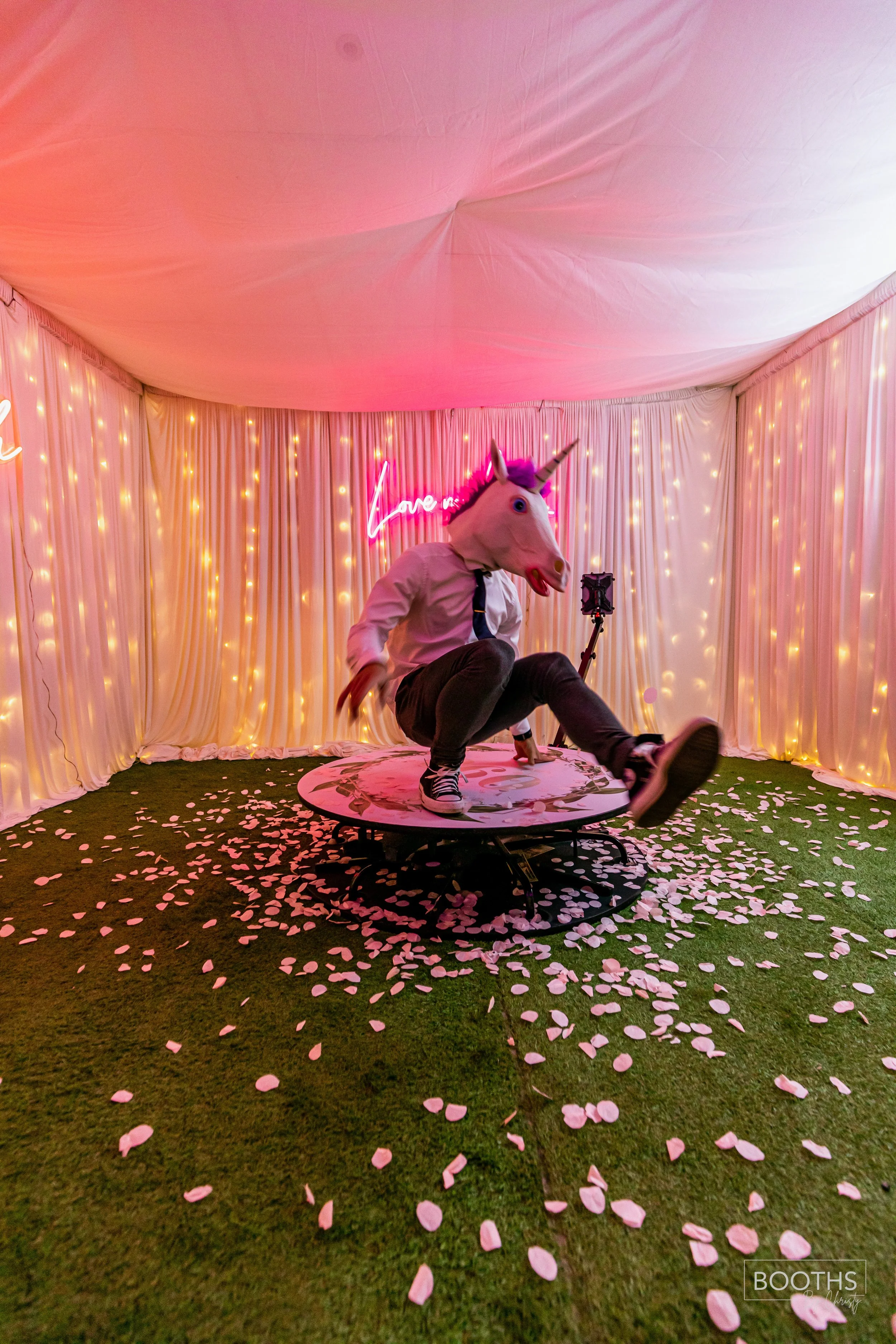 Person wearing a unicorn mask, white shirt, black tie, and black pants dancing on a round platform amidst pink flower petals and string lights in a decorated tent with a pink neon sign that says 'Love'.