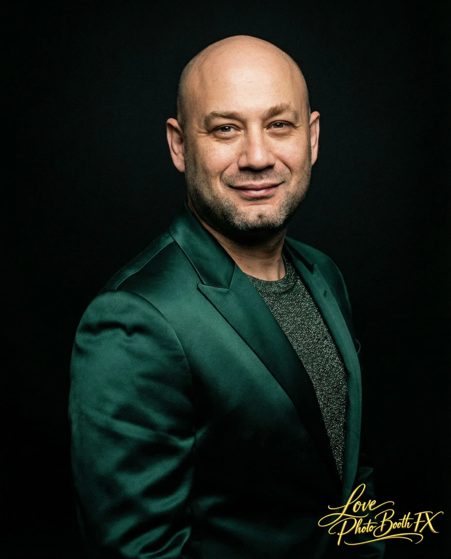 A smiling man with a bald head and beard, wearing a dark green blazer over a shimmering shirt, against a black background, with a gold logo in the bottom right corner that reads 'Love Photo Booth FX.'