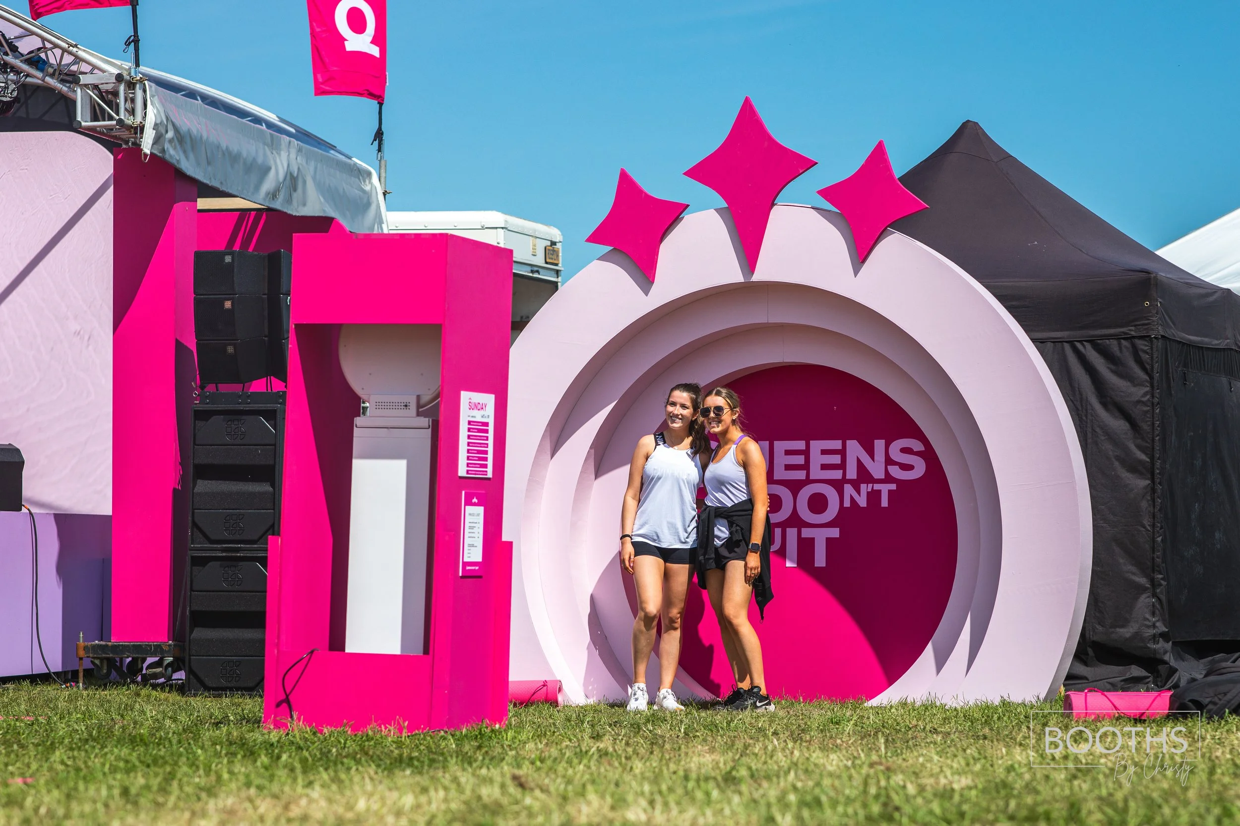 Two women standing in front of a pink and white booth with the partial phrase "QUEENS DON'T QUIT" on it at an outdoor event. The women are smiling, wearing athletic clothing, and sunglasses. The sky is clear and blue.