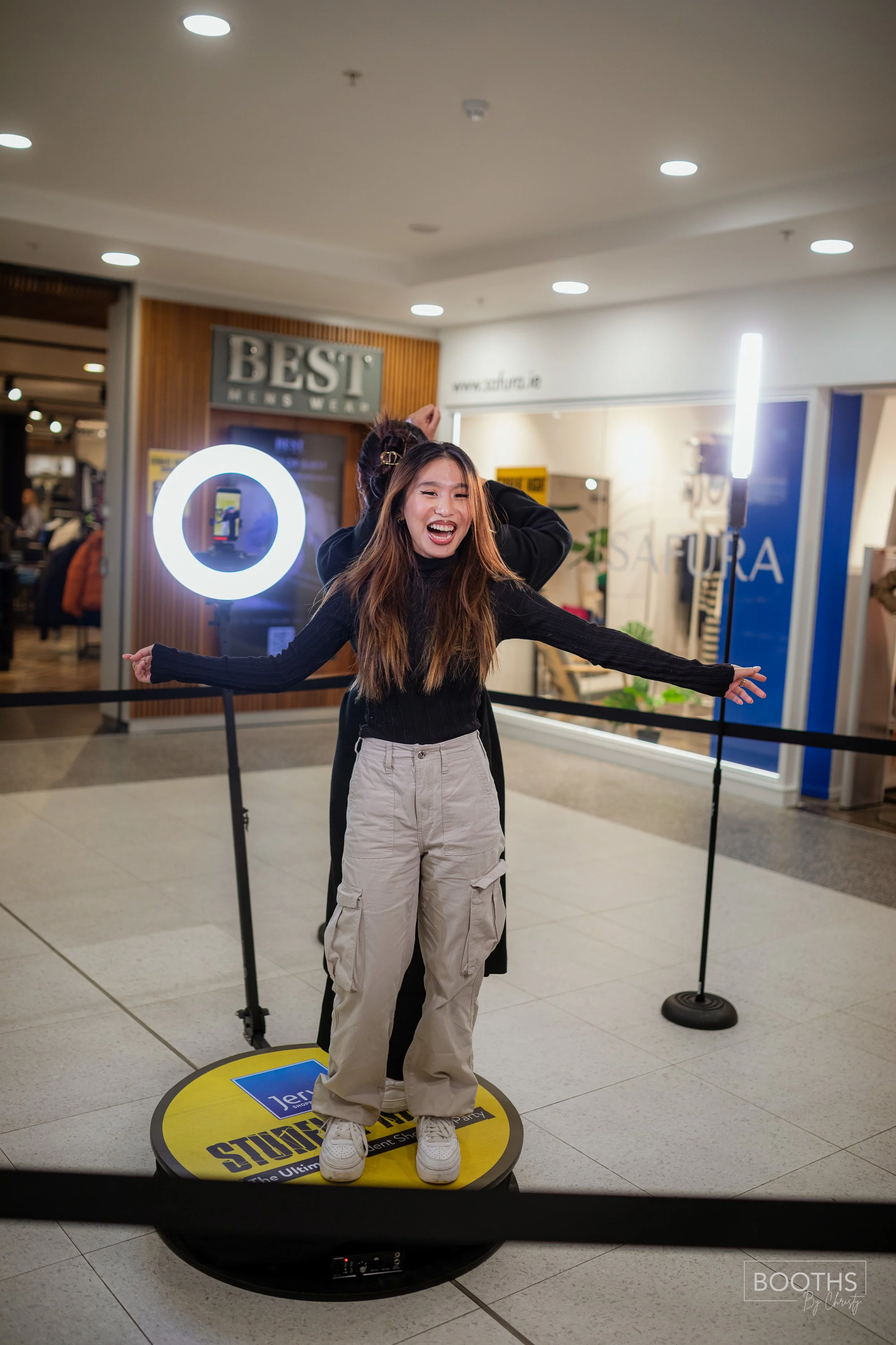 A woman standing on a round platform inside a shopping mall, smiling and celebrating, with a man hugging her from behind. There are bright lights and store signs in the background.