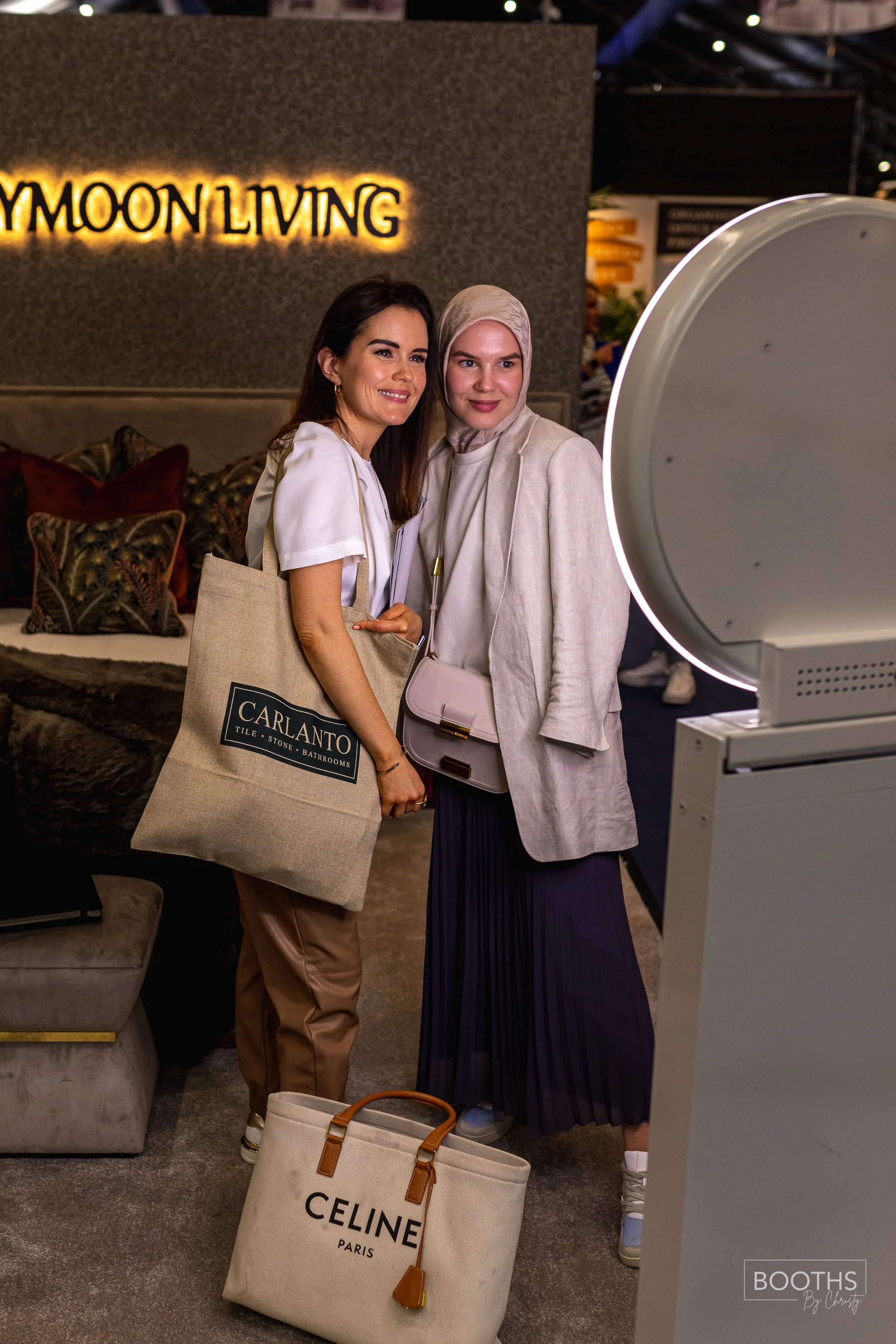 Two women taking a selfie at a booth with a large mirror, with a sign that says 'MOON LIVING' in the background. One woman is carrying a large beige tote bag with the label "CARLANTO" and the other has a large Céline bag on the floor. The scene appea