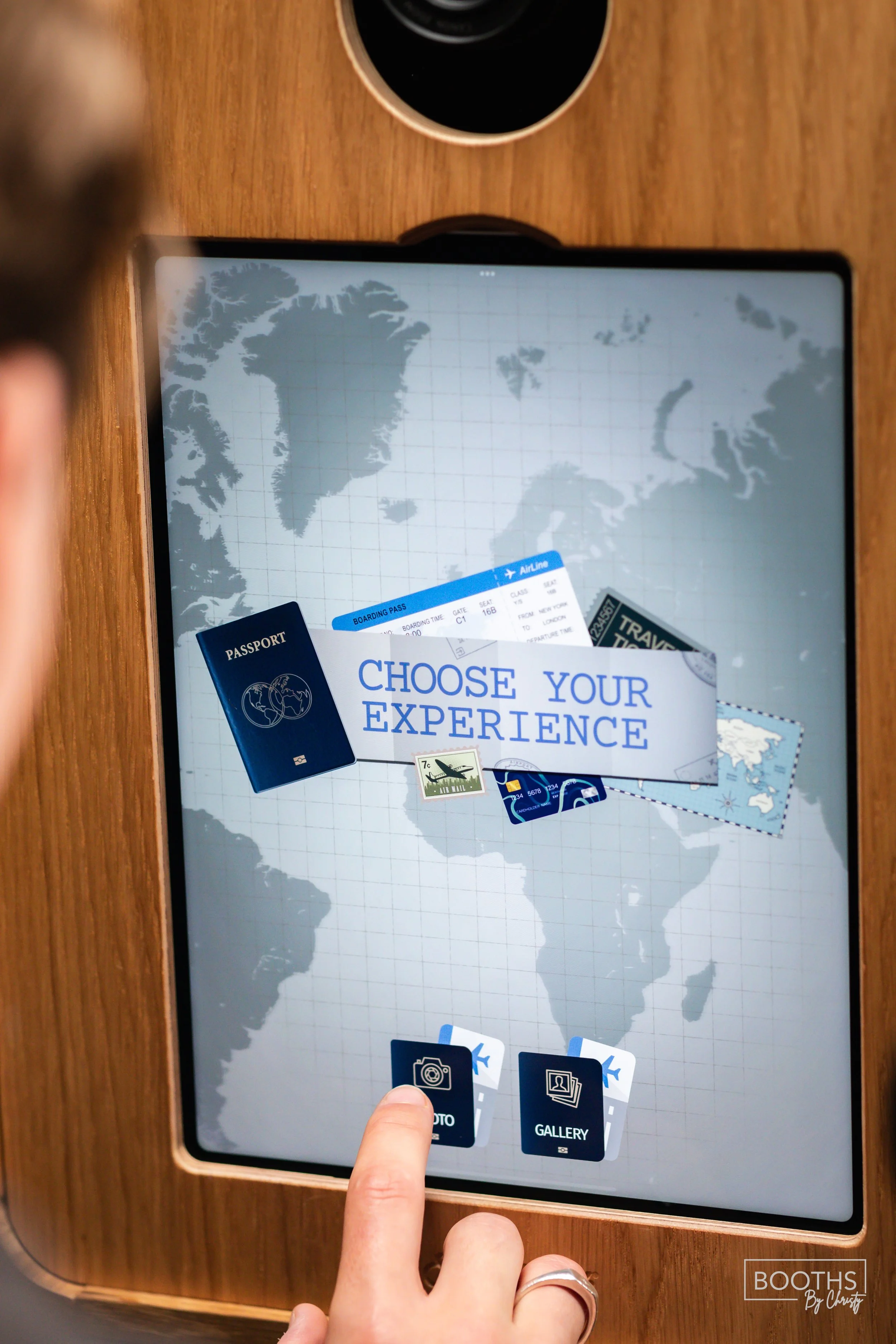 A person's finger is touching a tablet screen with travel options, including a passport, boarding passes, and travel stamps, on a wooden surface, with a world map background and a cup of coffee nearby.
