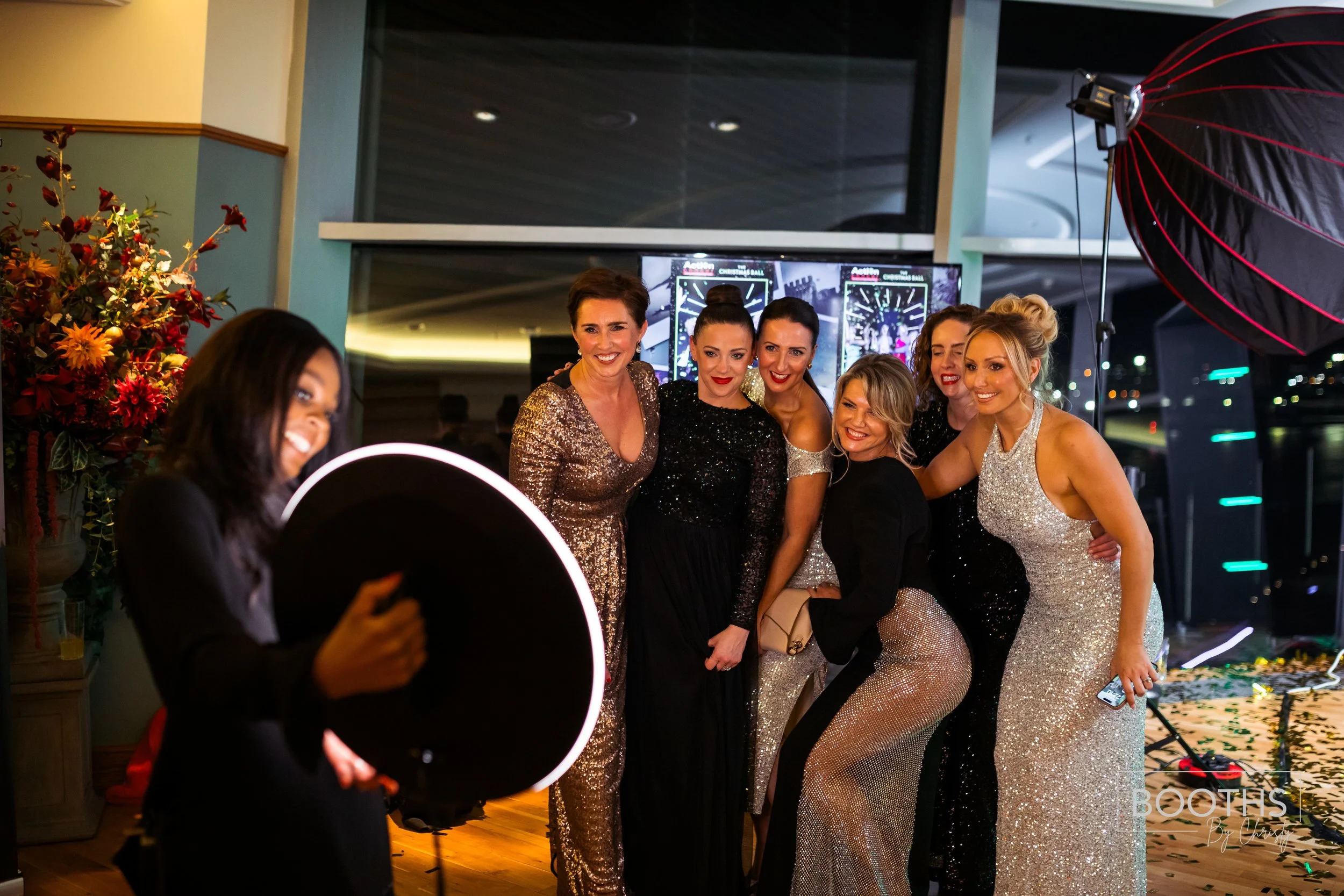 Group of women dressed in glamorous evening gowns taking a group photo at a party with a photographer and professional lighting setup.