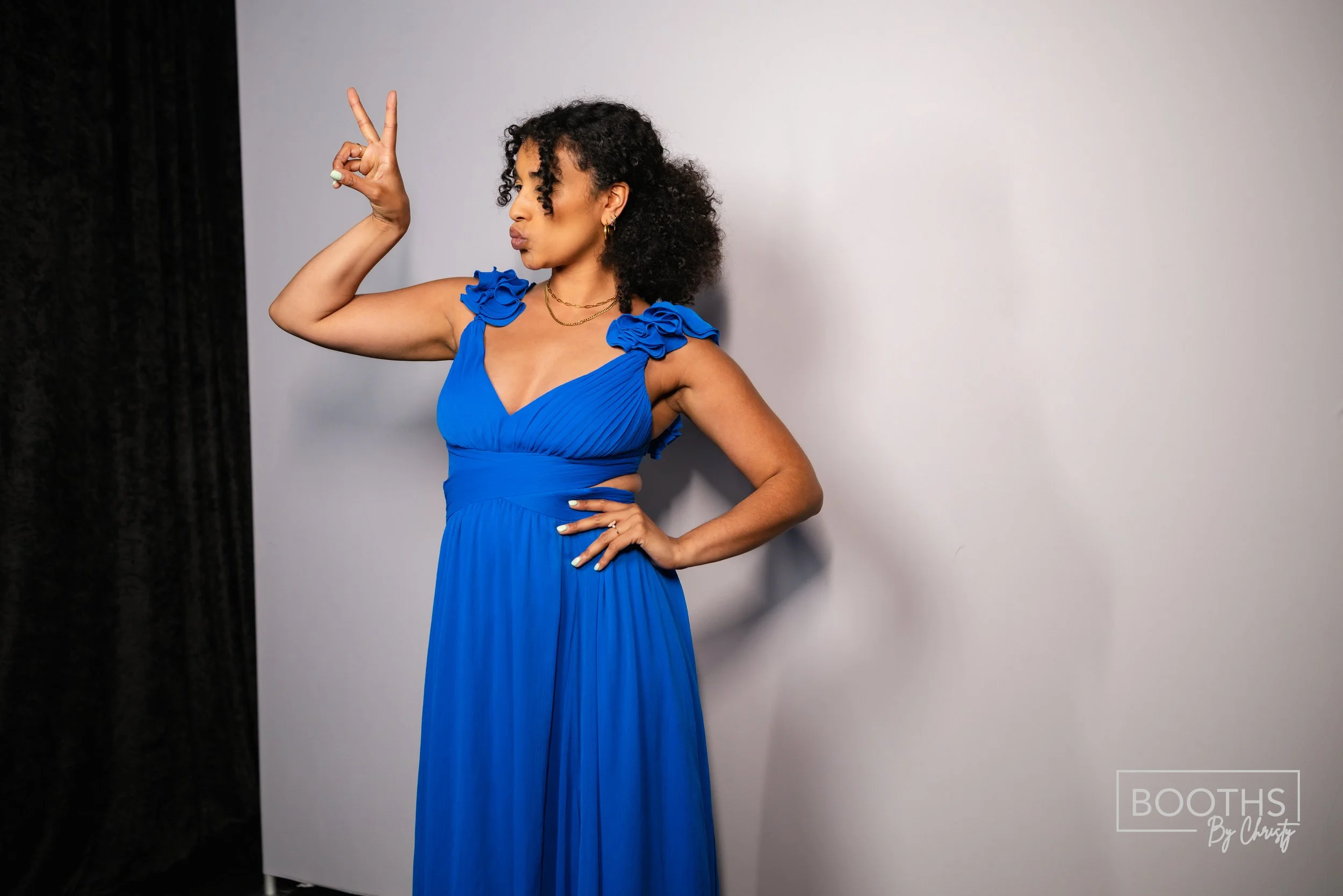 A woman in a bright blue dress with ruffles on the shoulders standing against a light gray backdrop, making a peace sign with her right hand and resting her left hand on her hip.