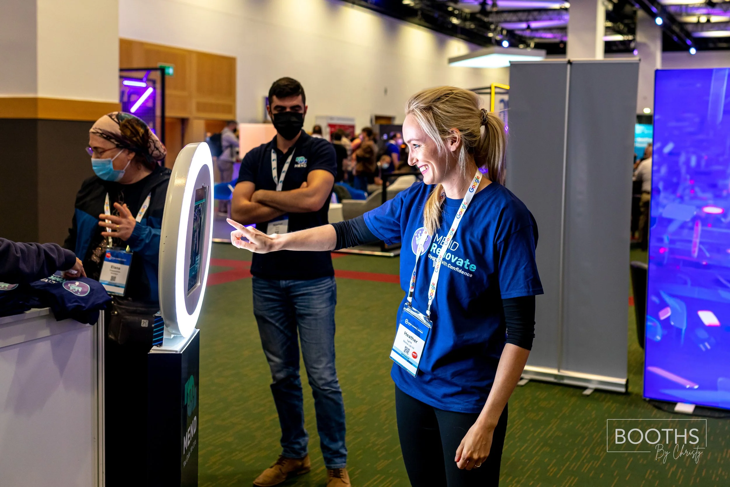 A woman with blonde hair smiling and taking a selfie with her smartphone at a tech event, standing next to a digital kiosk. Two men and a woman wearing masks are visible in the background, with a large screen and other event booths around.