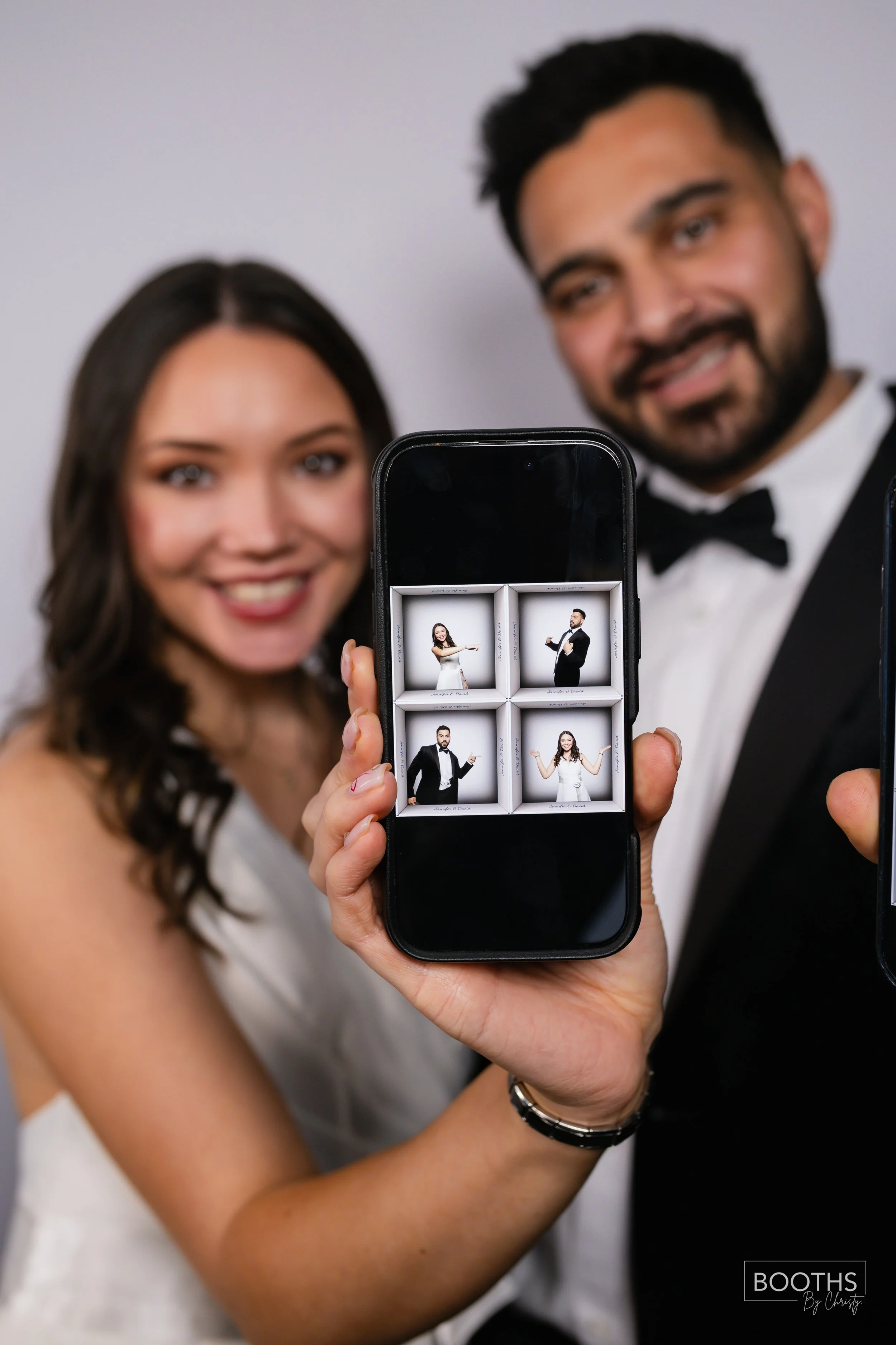 A woman and a man dressed in formal wedding attire taking a selfie while holding a photo booth strip showing four pictures of themselves. The woman has long curly hair and the man has a beard and mustache.