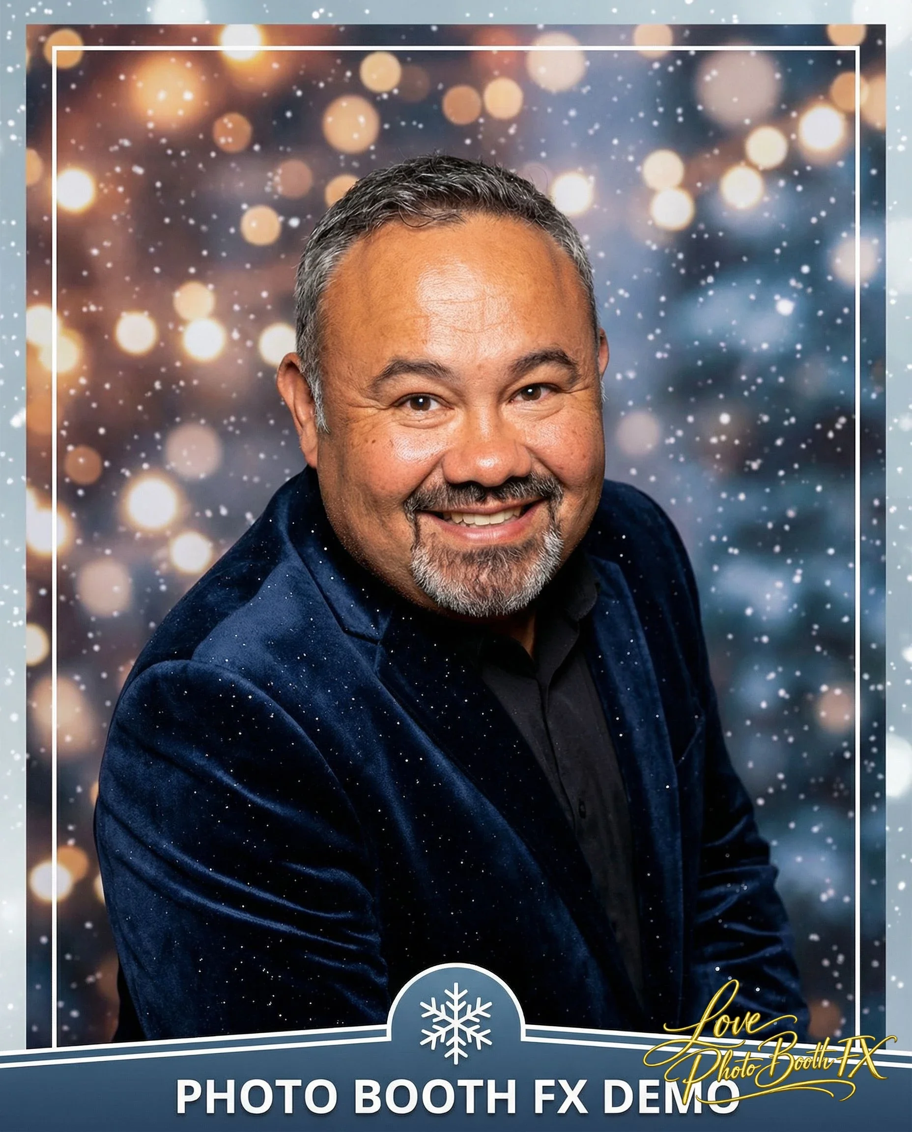 A smiling man with short gray hair and a beard wearing a dark blue velvet blazer with a black shirt against a festive, blurred Christmas tree with lights and snowflake decorations background.