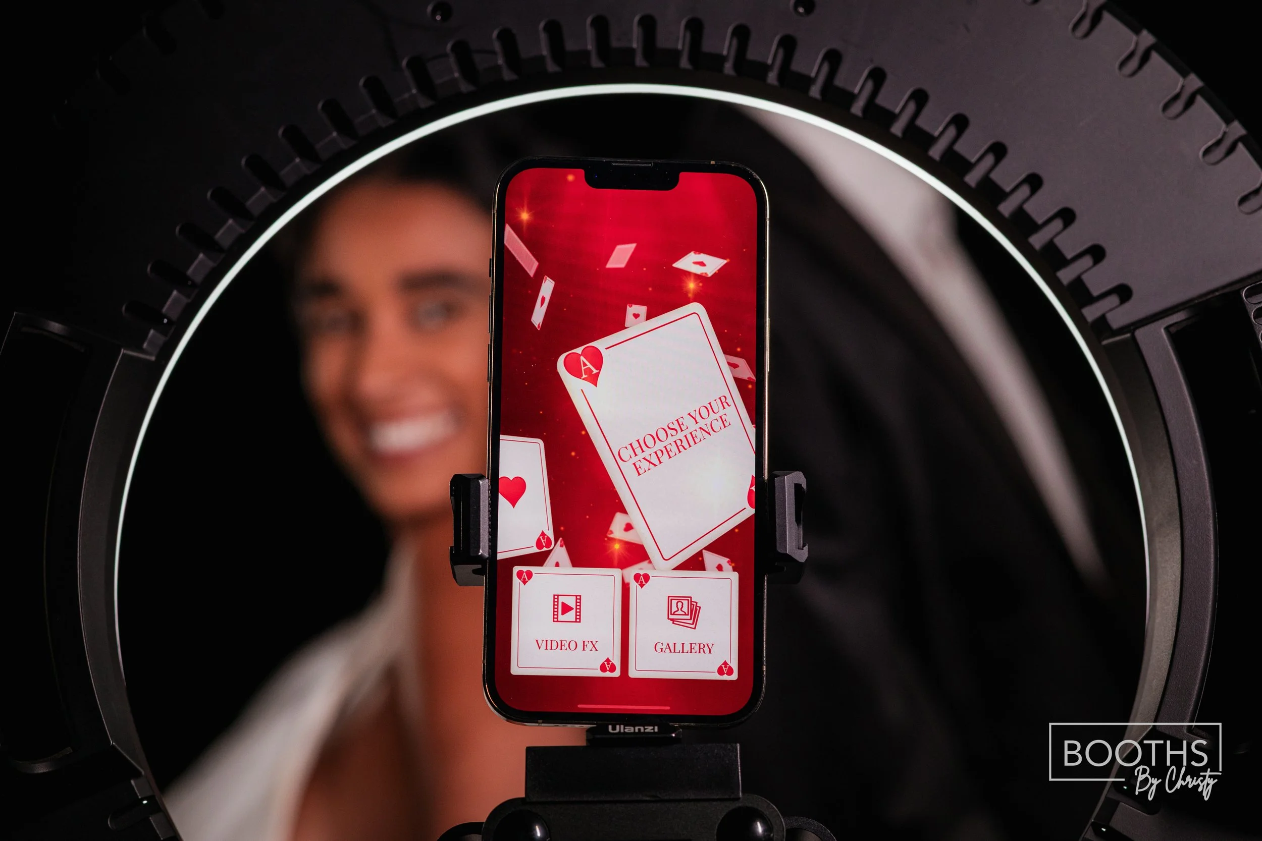 A smartphone mounted on a stand displaying a game menu with the text 'Choose Your Experience' and options for 'Video FX' and 'Gallery', in front of a smiling woman blurred in the background, inside a photobooth with a ring light.