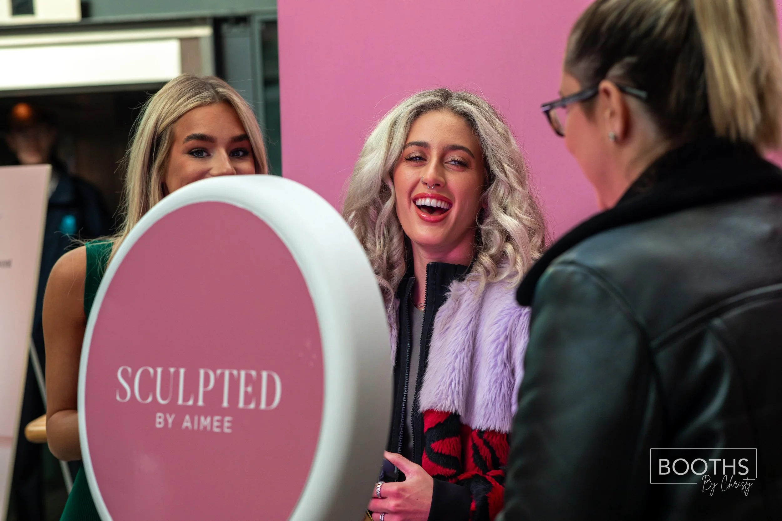 Two women smiling and talking at a booth, with a sign that reads 'SCULPTED BY AIMEE' in front of them. One woman has blonde hair and is wearing a black and red jacket, and the other has gray curly hair and is wearing glasses and a black leather jacke